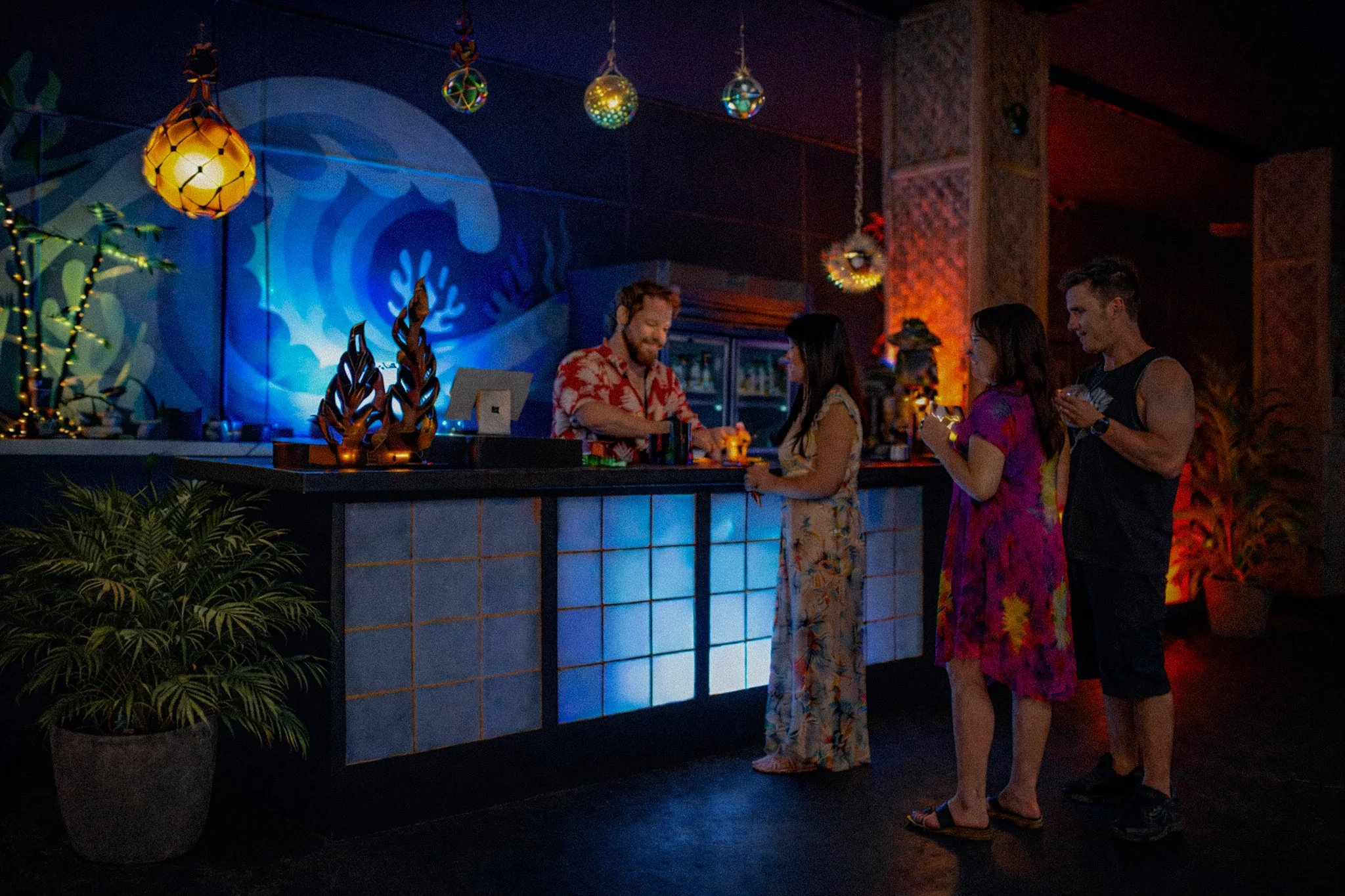Bartender prepares a tropical cocktail for guests in a tiki bar, featuring ocean and volcano themed areas.
