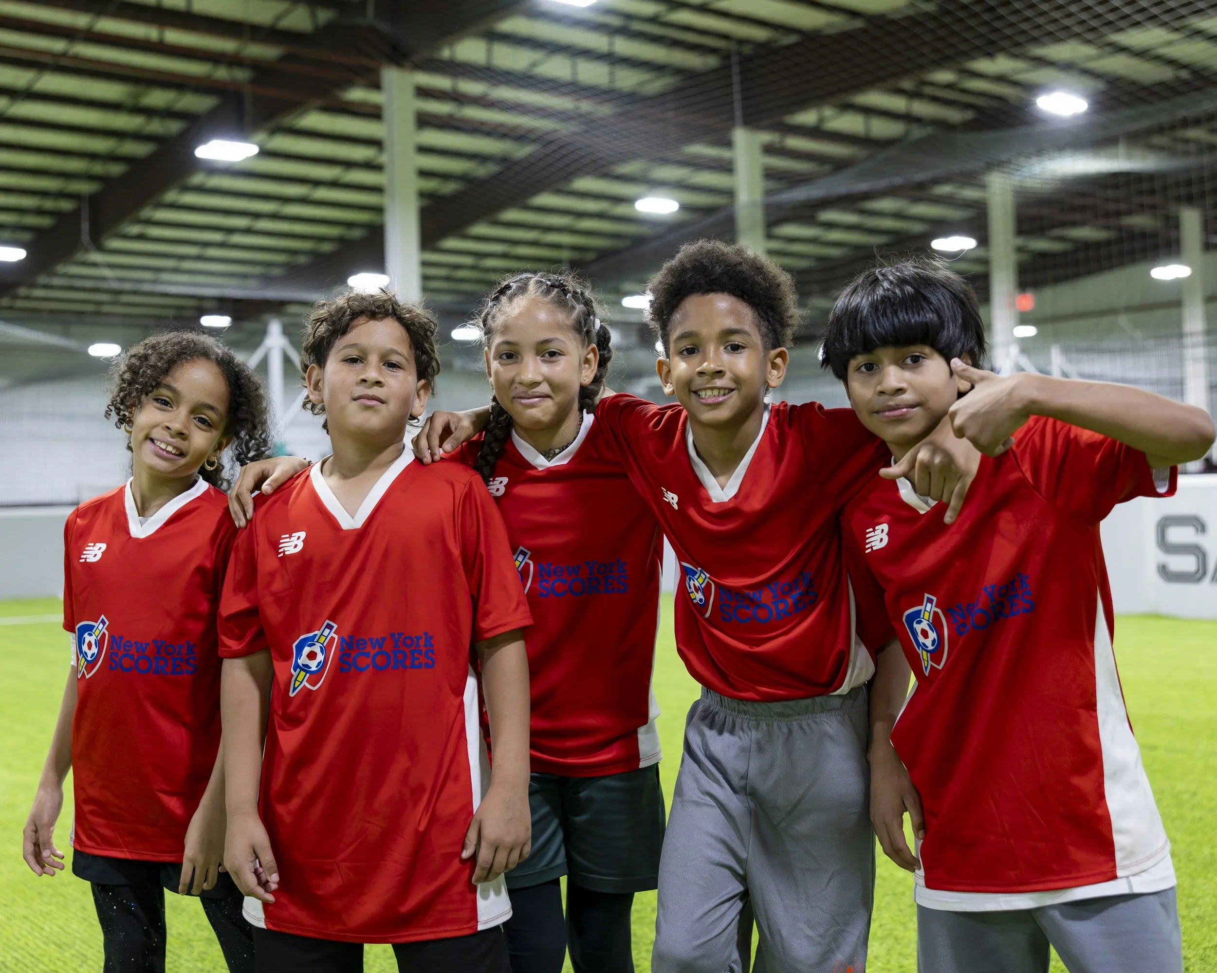 Group of five young kids in red soccer jerseys standing inside an indoor soccer facility on a green field, smiling and posing for a photo.