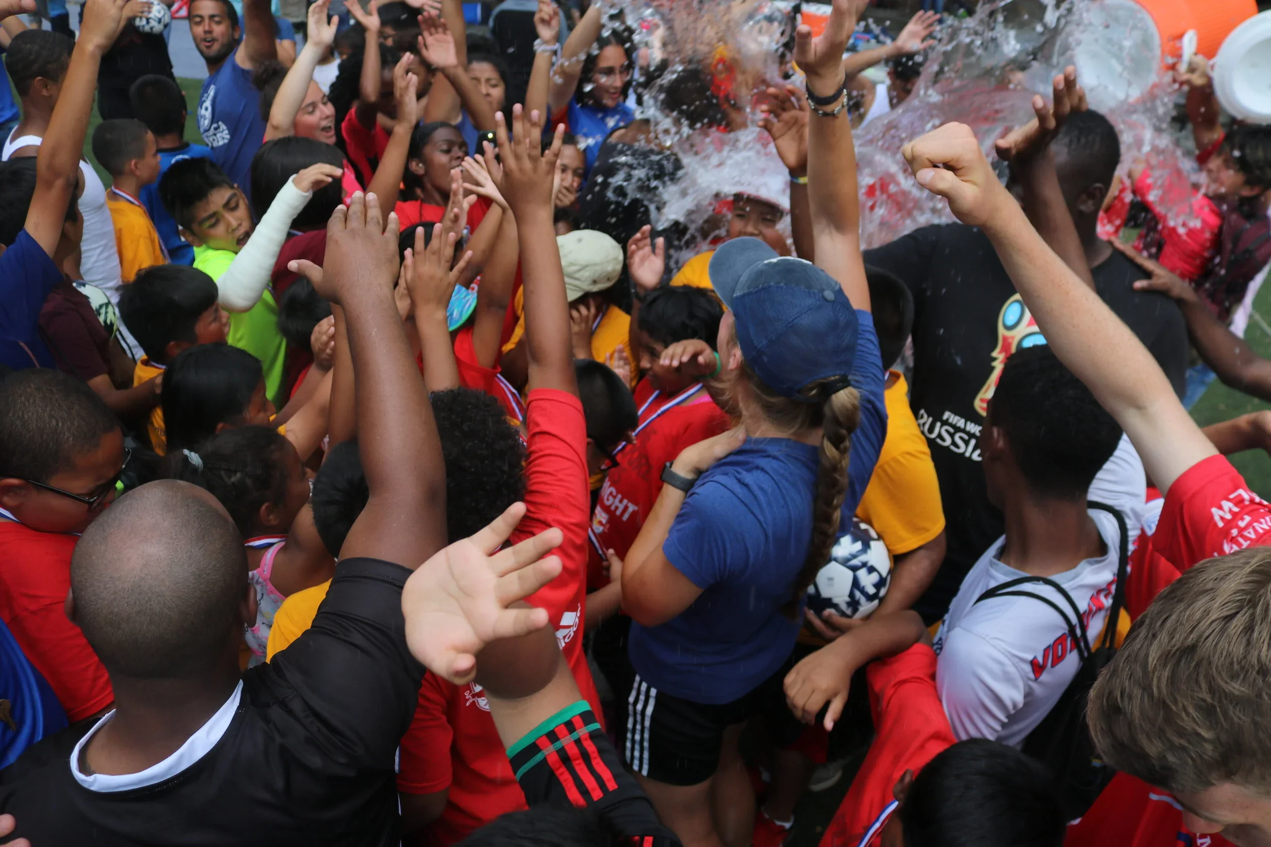 Crowd of children and adults celebrating in the rain, some wearing soccer jerseys, in a festive outdoor event with water splashing.