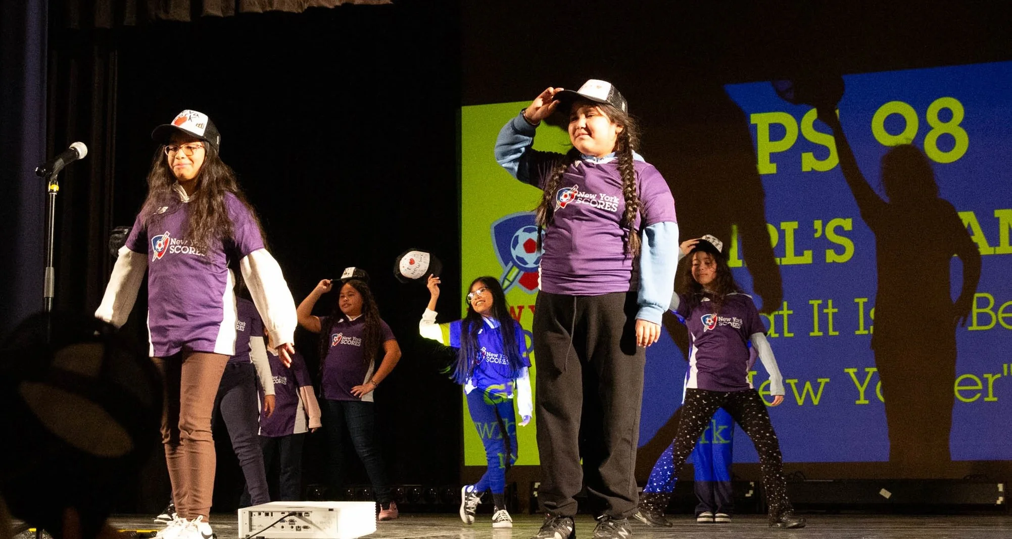 Group of children performing on stage, wearing purple shirts with 'New York Scores' logo, some wearing hats, with a microphone stand and a large screen in the background.