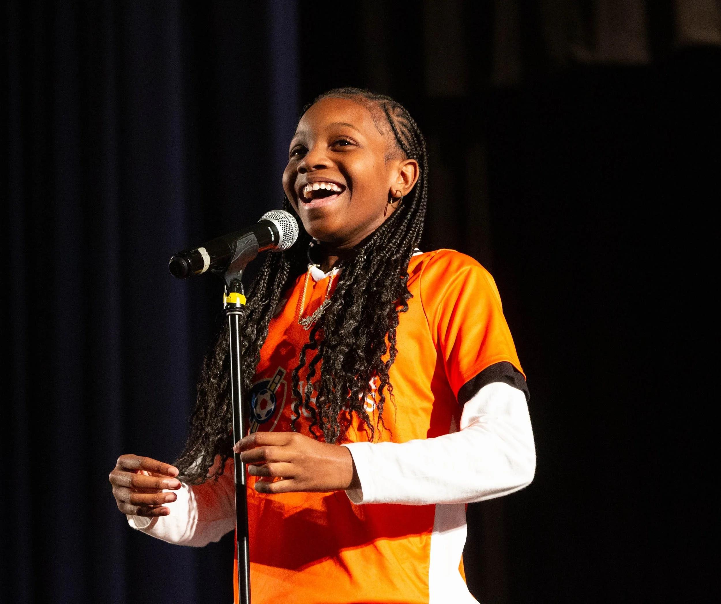 A young girl with braided hair, wearing an orange sports jersey over a white long-sleeve shirt, smiling and speaking into a microphone on stage.