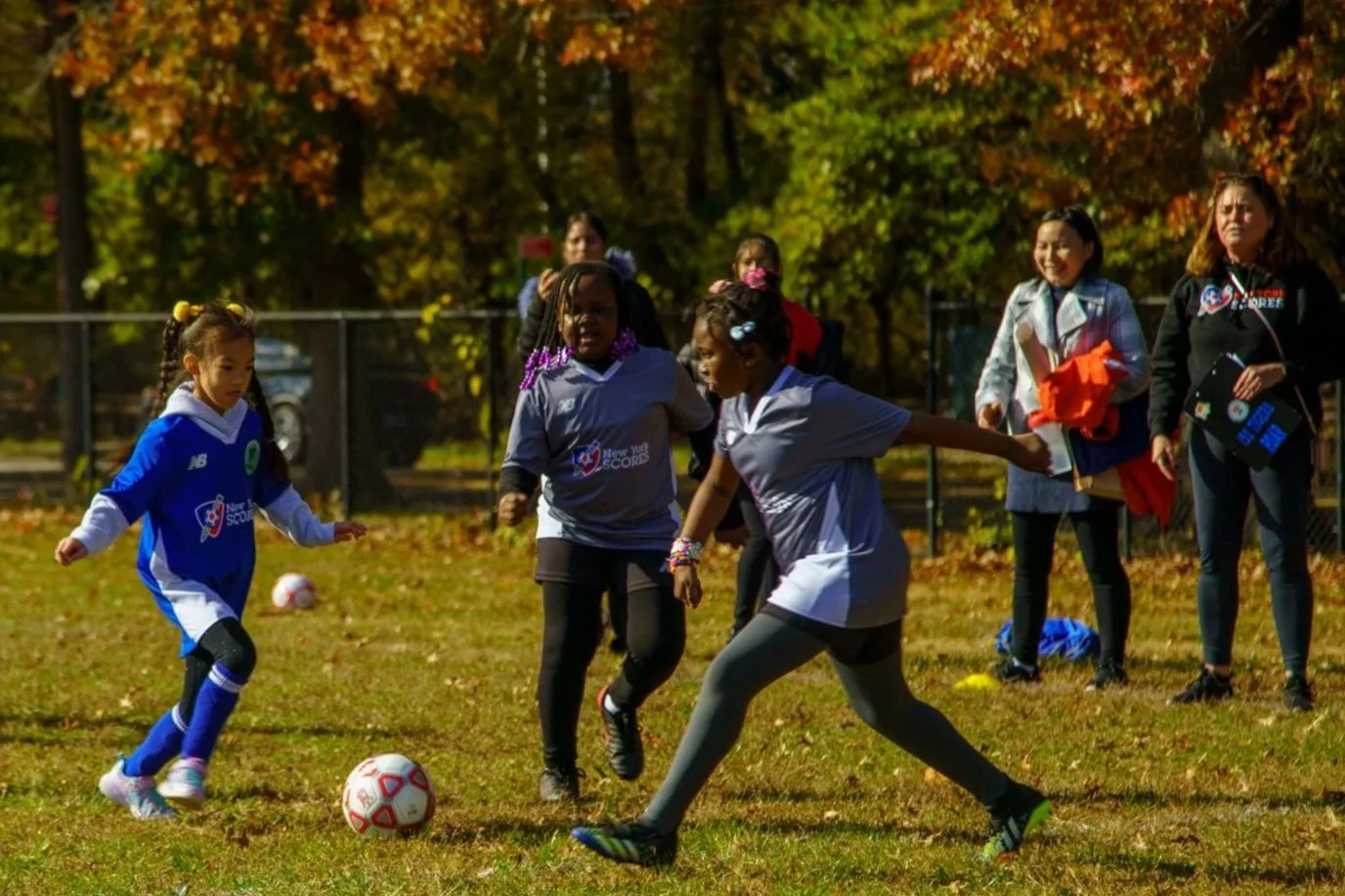 Children playing soccer outdoors during autumn, with colorful fall foliage in the background.