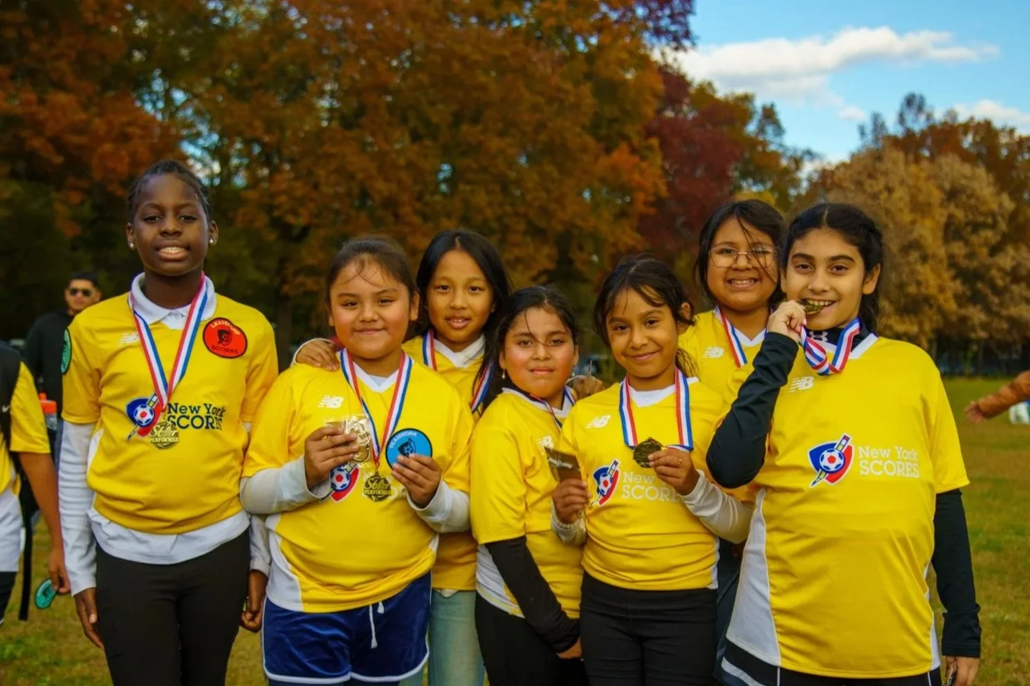 Group of young girls in yellow soccer jerseys with medals around their necks, smiling and holding medals and chocolates, outdoors with autumn trees in the background.