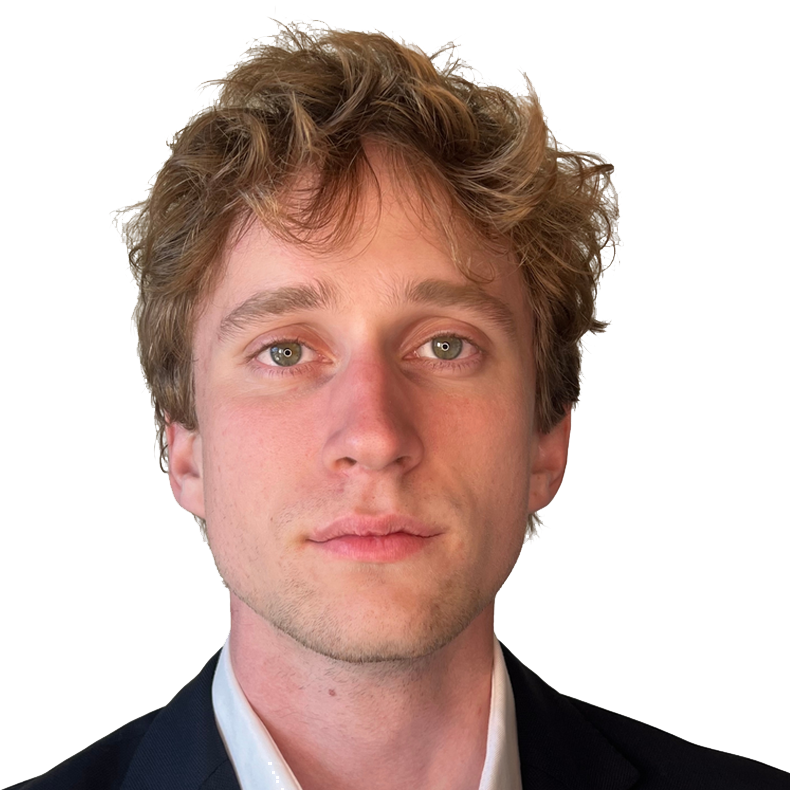 Close-up of a young man with light brown, curly hair and blue eyes, wearing a dark blazer and white shirt, against a plain background.
