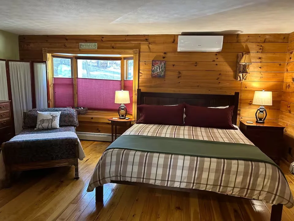 Bedroom with wood-paneled walls, a large bed with striped bedding and maroon pillows, two bedside lamps, a chaise lounge with a decorative pillow, a window with red shades, and a small sign above the window.