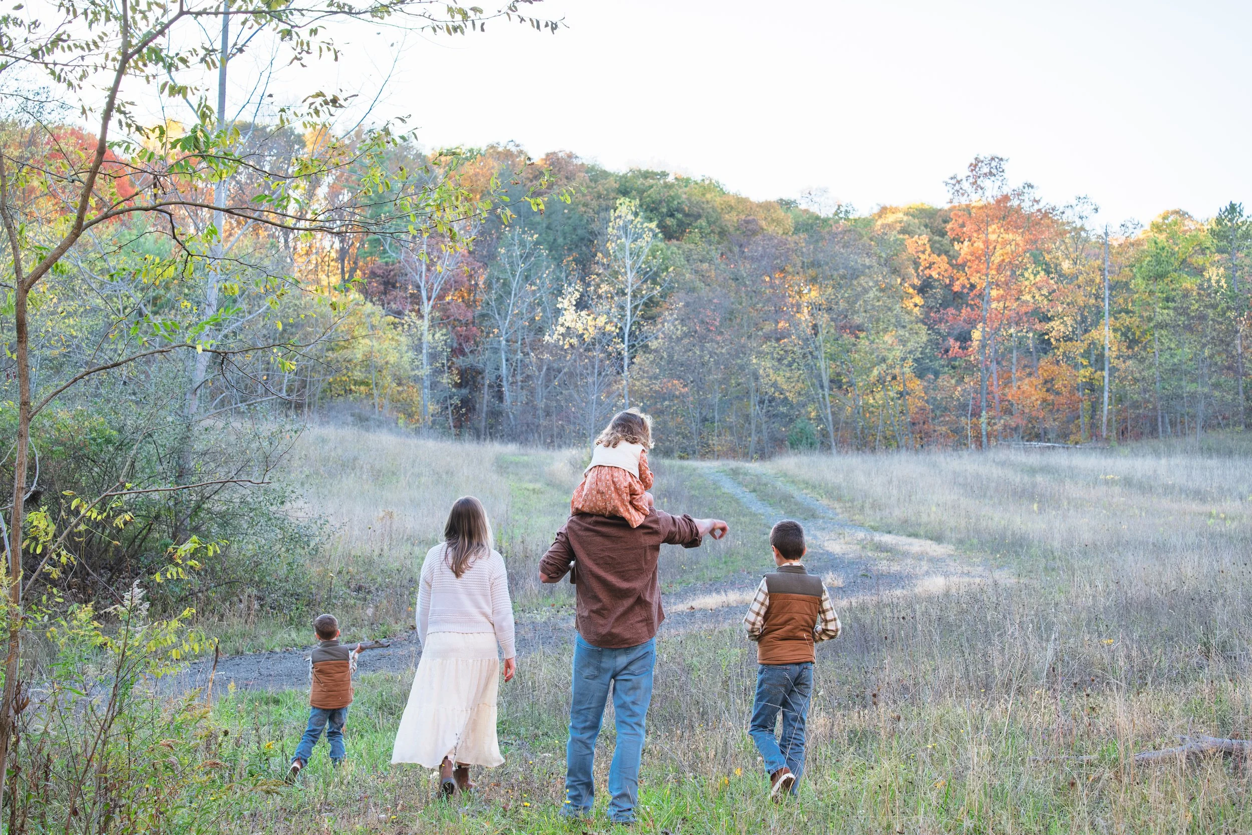 A group of children and an adult walking toward a wooded area with fall foliage, on a trail in a grassy clearing.
