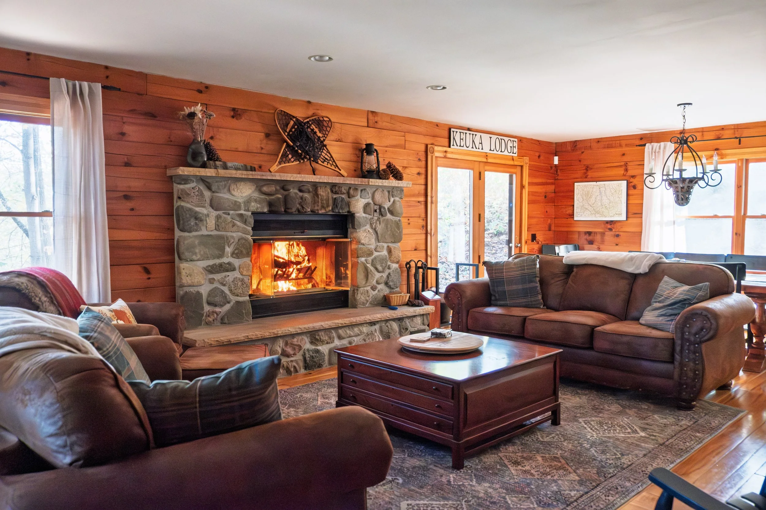 Living room with a stone fireplace, a fire burning, wooden walls, and large windows with curtains. There are sofas with plaid cushions, a wooden coffee table, and a chandelier.