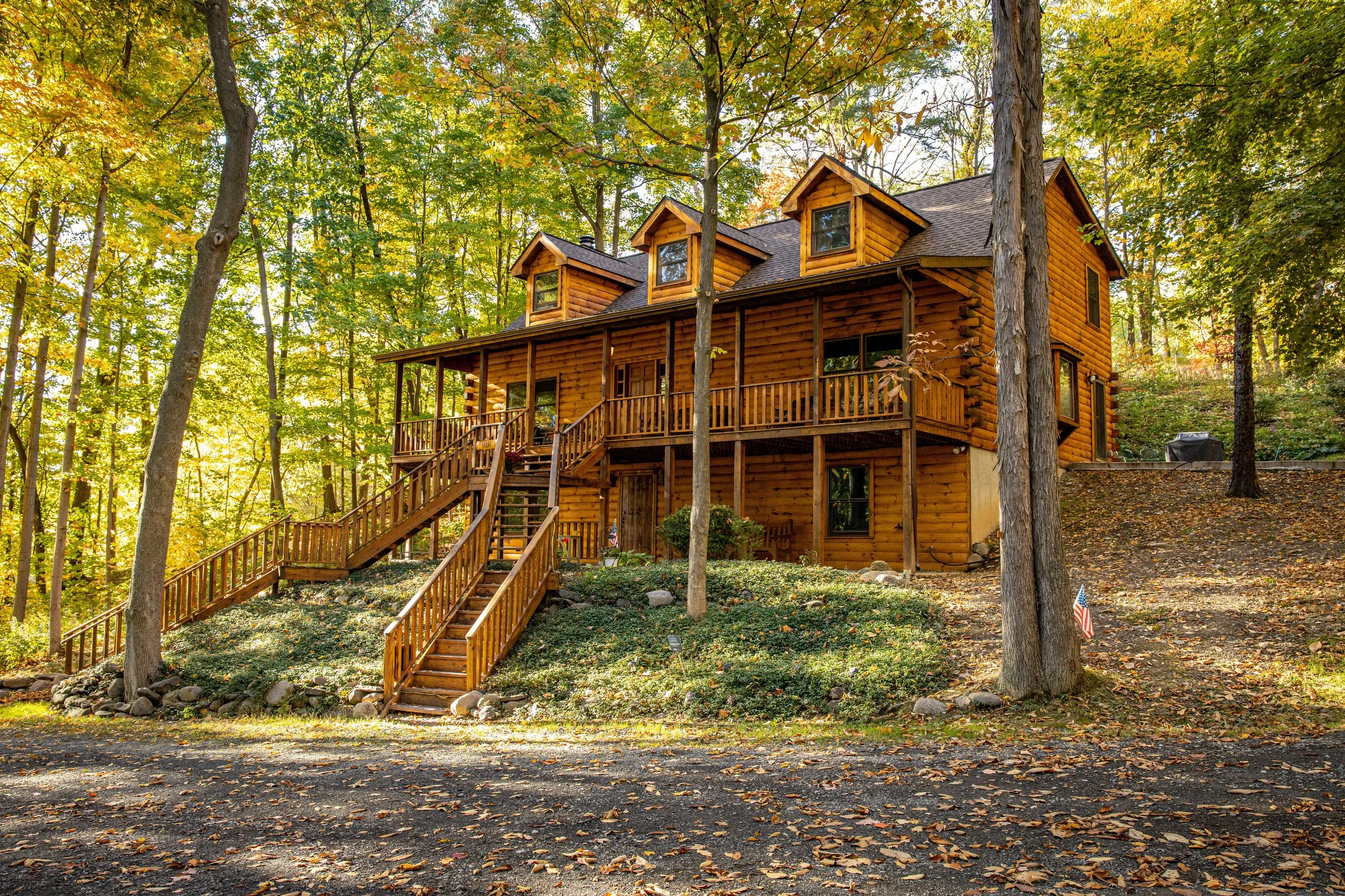 A two-story wooden house in a wooded area with trees displaying fall foliage. The house has a front porch with stairs and a railing, and multiple dormer windows on the roof. Fall leaves cover the ground, and sunlight filters through the trees.