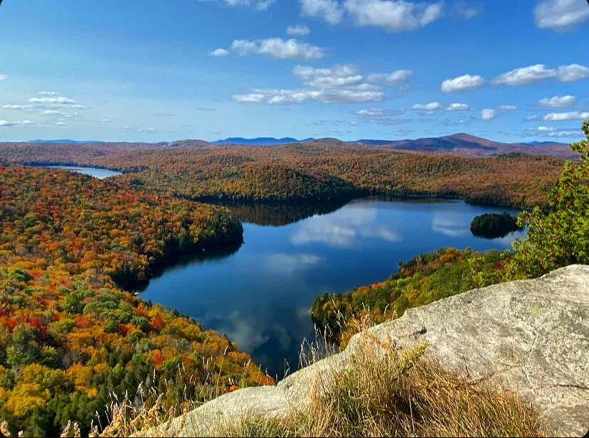 A scenic view of a lake surrounded by colorful autumn trees and rolling hills under a partly cloudy sky, taken from a high vantage point with a rocky ledge in the foreground. Gorgeous Vermont fall foliage scenery.