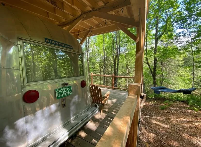 A vintage 1965 Airstream, under roof cover, and overlooking a flowing brook in the woods.