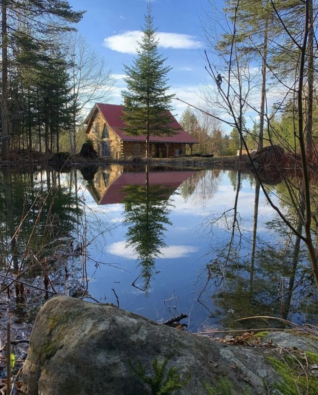 A quintessential Vermont log cabin with a red roof sits beside a calm and peaceful pond, reflecting the house and surrounding trees, with a cloudy blue sky overhead.