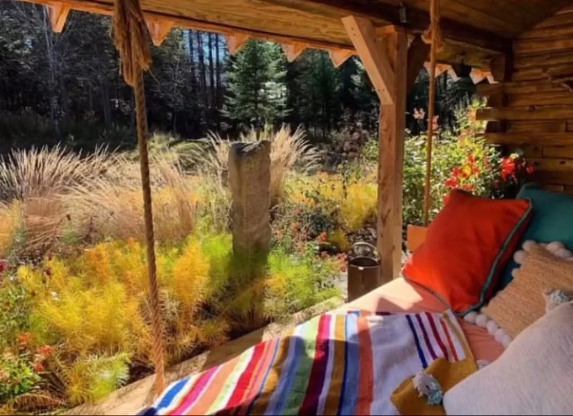 View from a cozy porch bed swing with colorful pillows and blanket, overlooking a peaceful garden with flowers and trees. A private and unique log cabin in the woods in Vermont for couples or small groups.