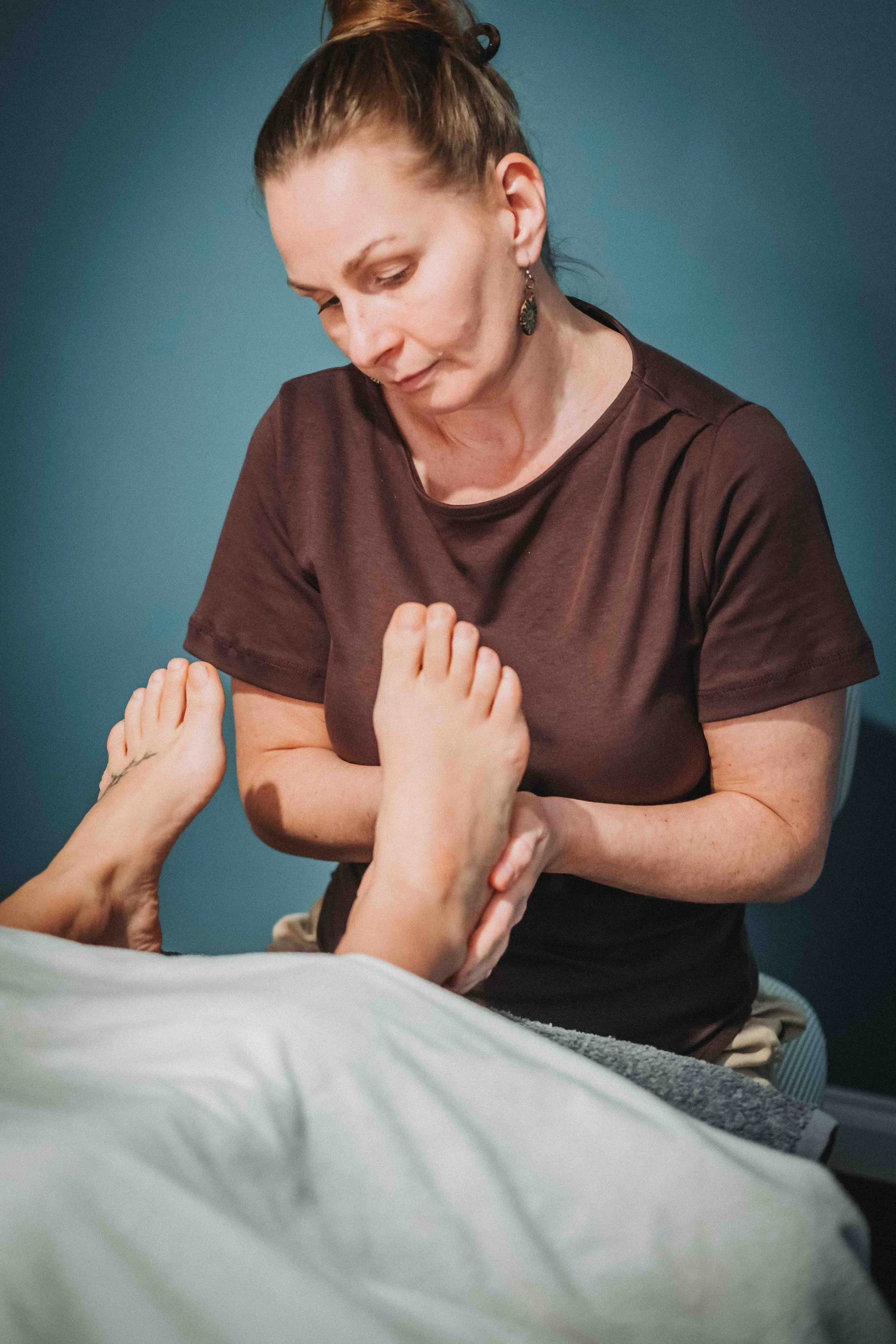 A woman with brown hair tied back, wearing a dark brown shirt and earrings, is sitting and looking down at her arm while holding a person's hand, which is gently touching her arm. The background is a plain blue wall.