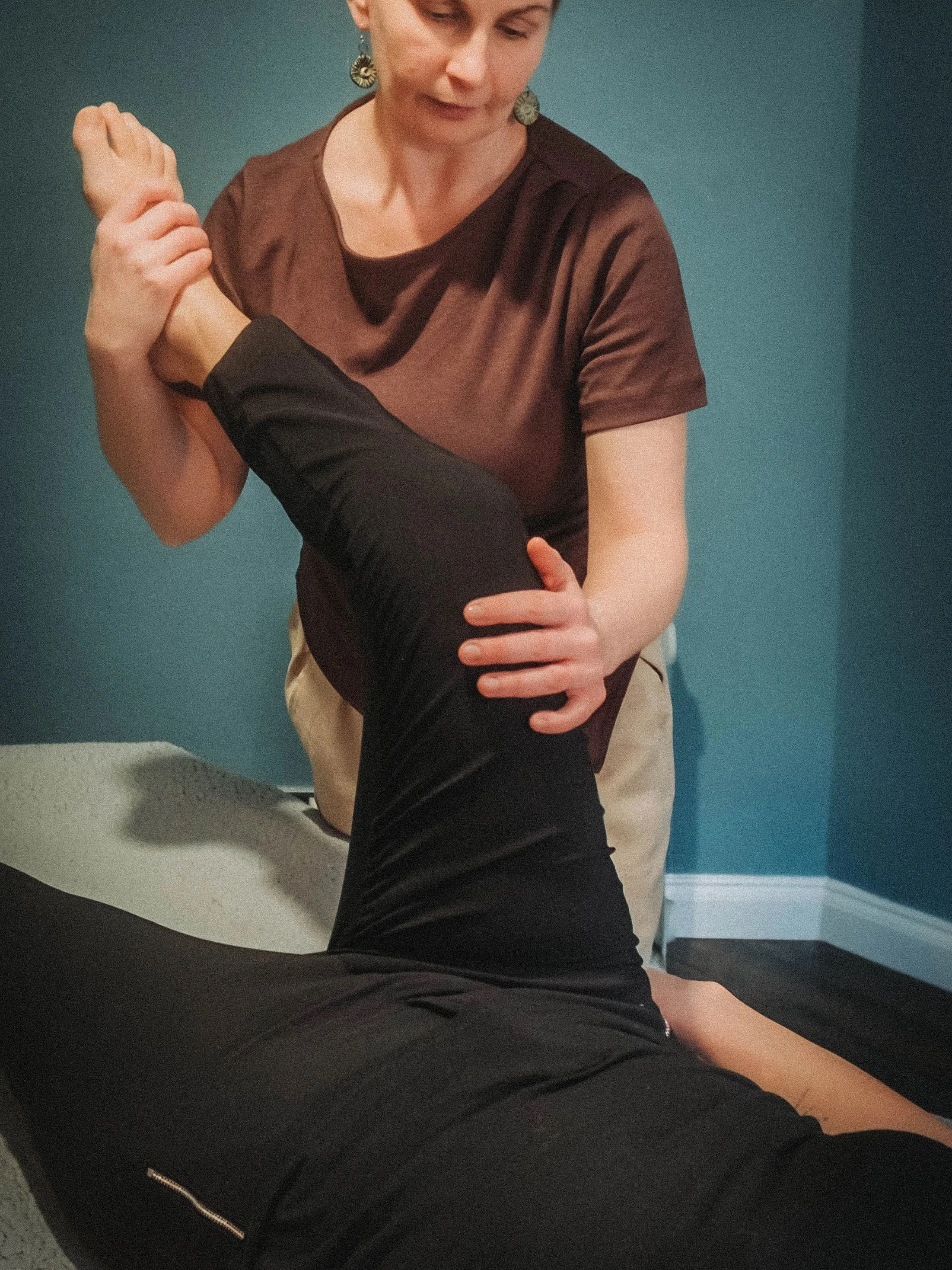 A woman wearing earrings and a brown shirt is performing a stretching exercise, holding her arm and elbow as she stretches.