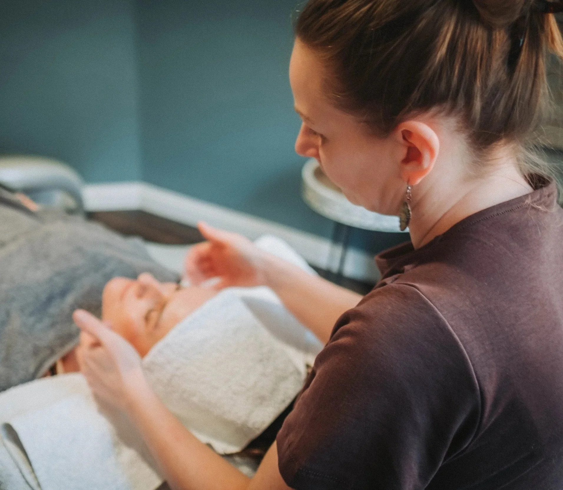 A woman with brown hair tied back, wearing earrings and a brown shirt, is seated and appears to be in a healthcare setting, possibly receiving a medical injection or vaccination from a healthcare worker.
