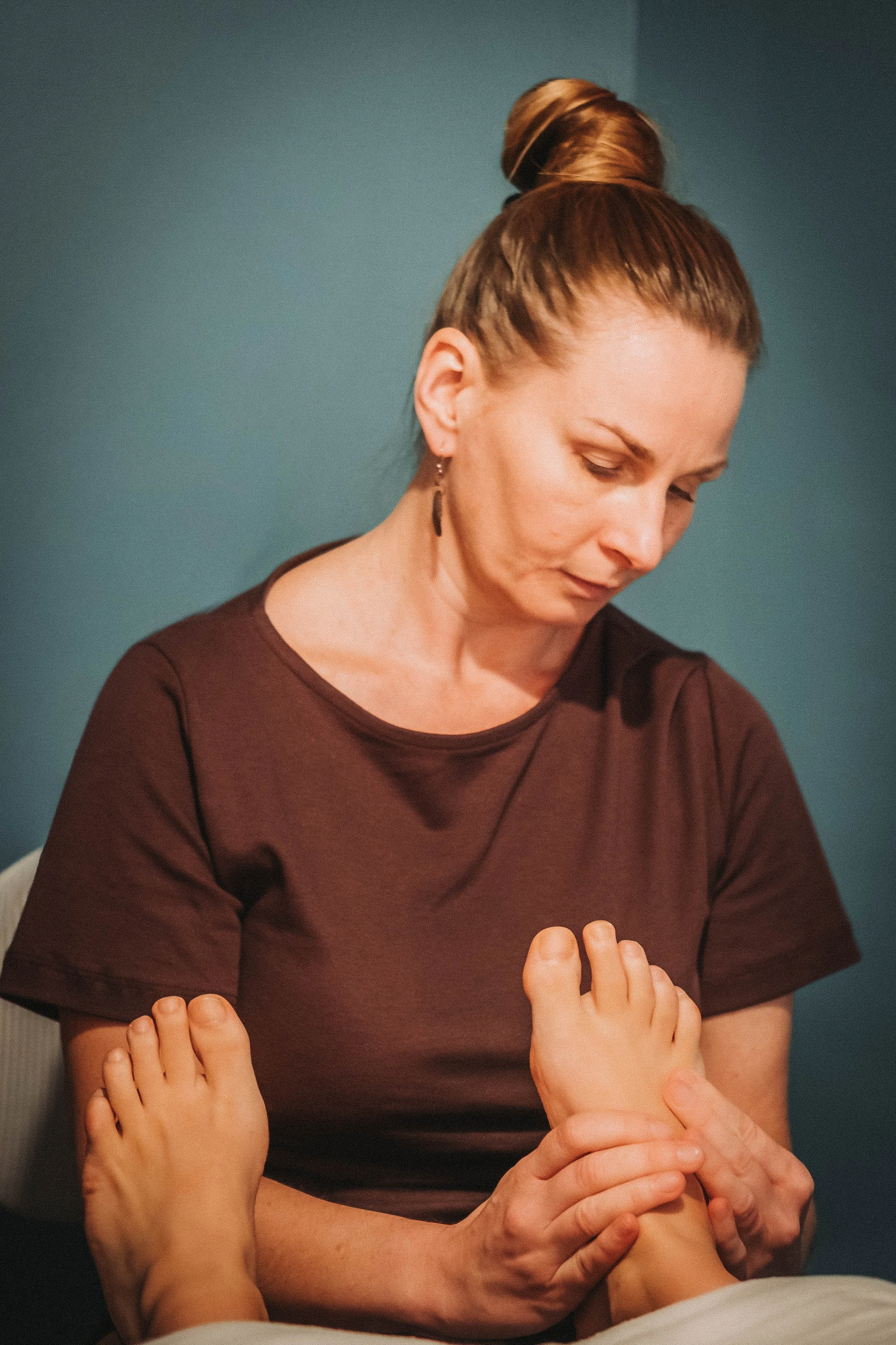 A woman receives a foot massage from another person. The woman has her hair tied up in a bun and is wearing a dark brown top. The massage is taking place in a setting with a teal wall background.