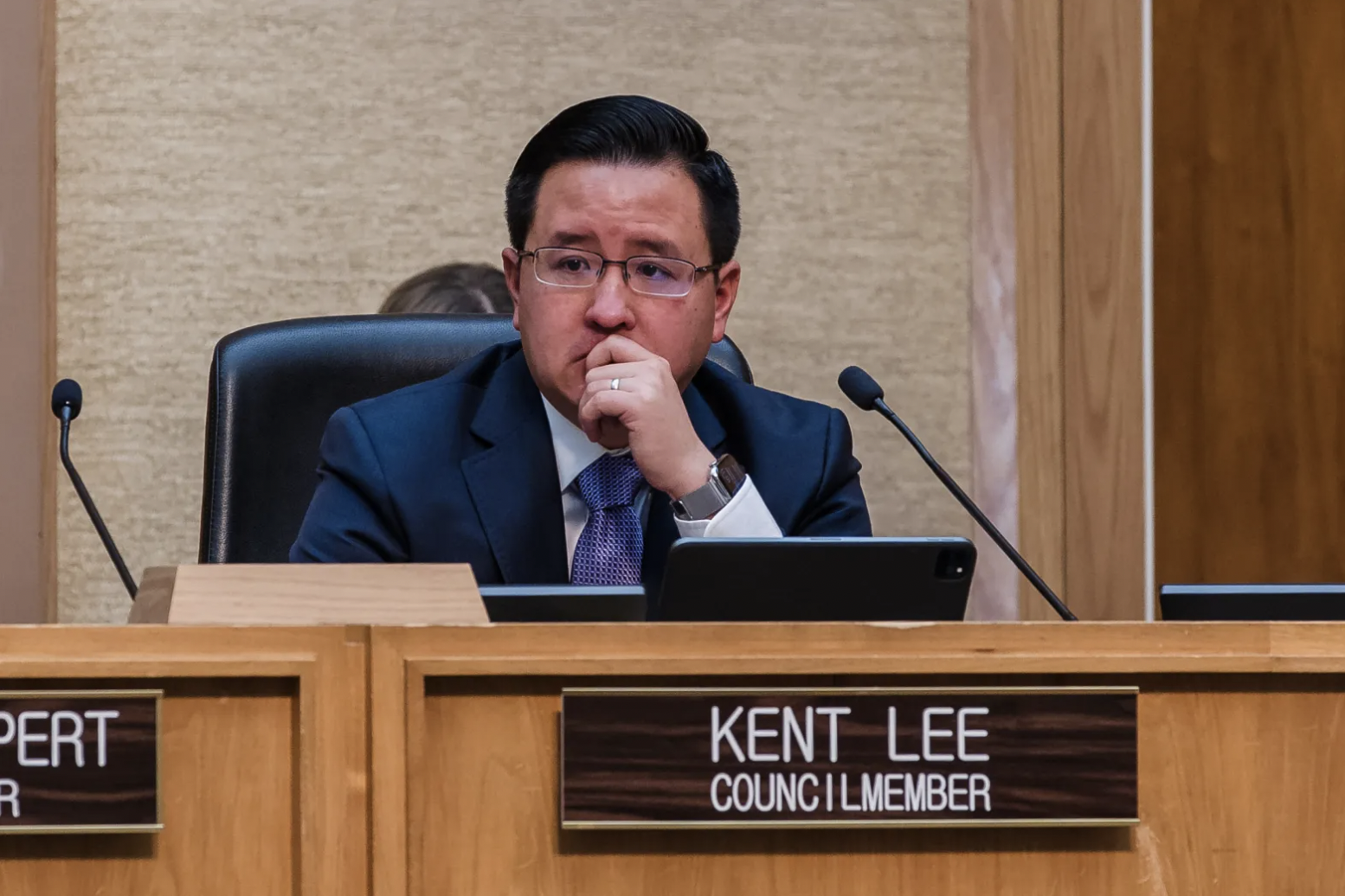 A man in a navy suit and glasses sitting at a wooden desk with a sign that reads 'KENT LEE COUNCIL MEMBER'. He is resting his chin on his hand, has a watch on his wrist, and is looking forward.