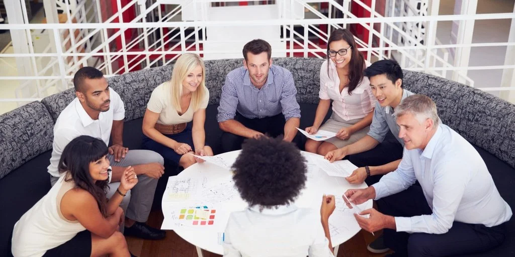 Group of diverse professionals sitting around a circular table, engaged in a discussion with papers and documents.