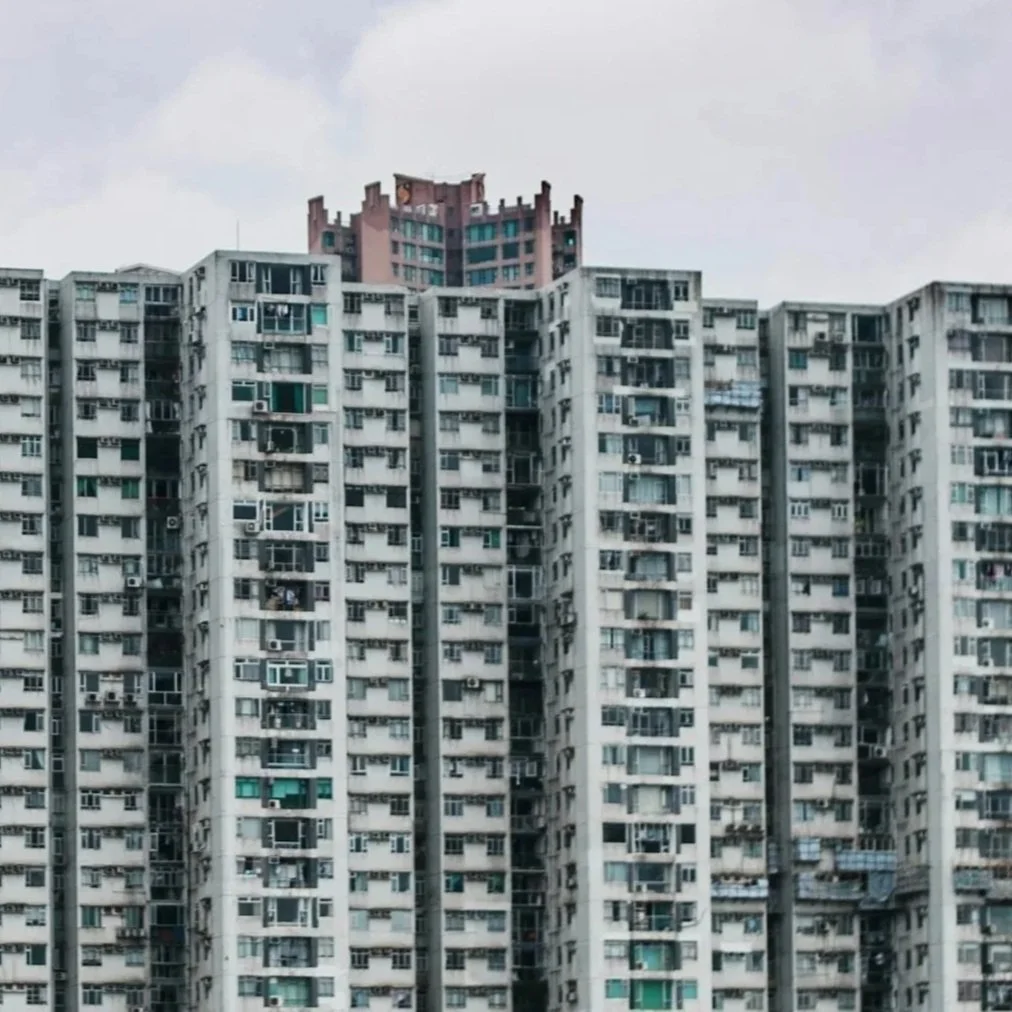 A tall, white apartment building with multiple windows and balconies, with another building towering over the density of apartments.