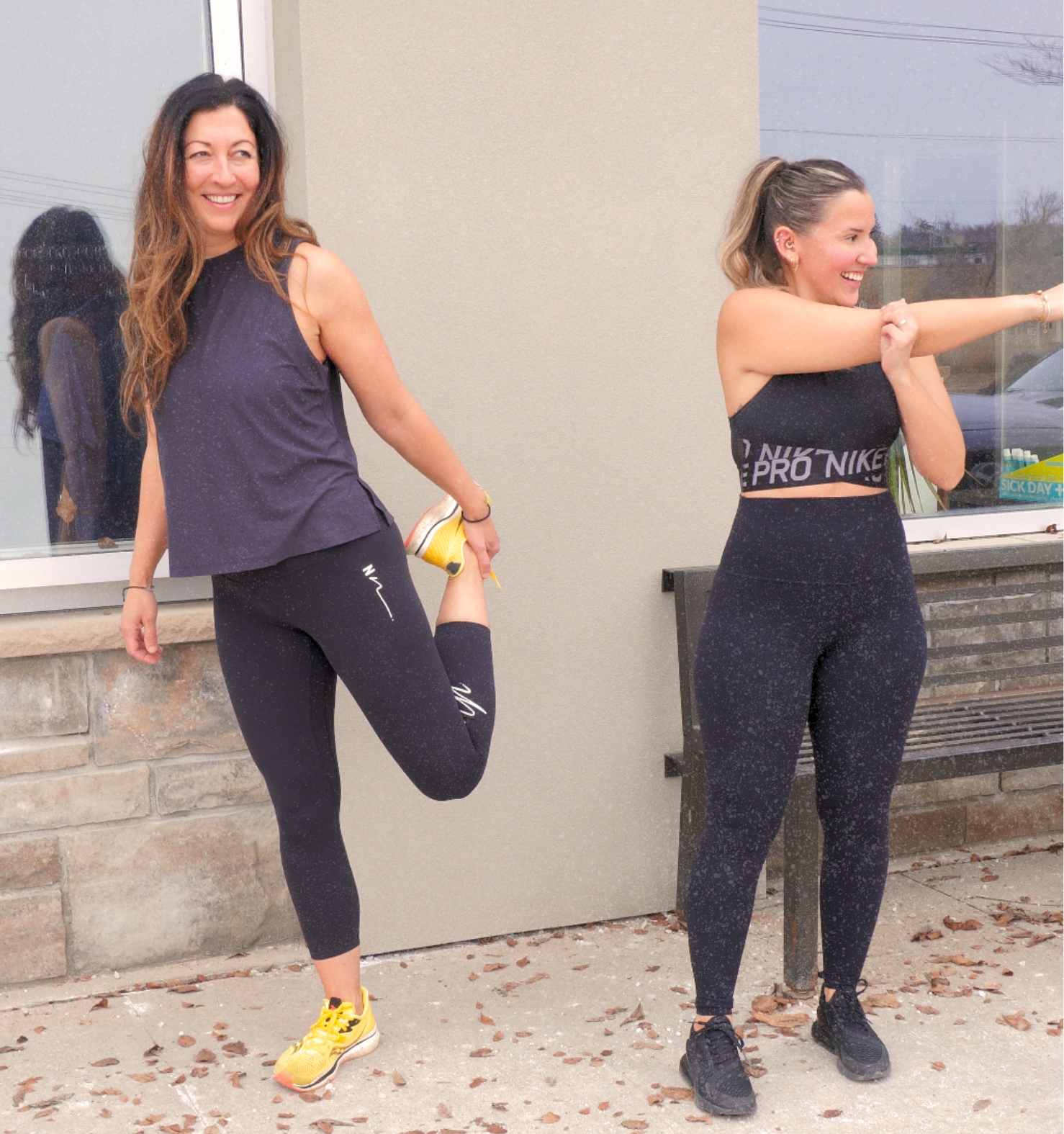 Two women in athletic clothing stretching outdoors, one holding her ankle behind her, both smiling.