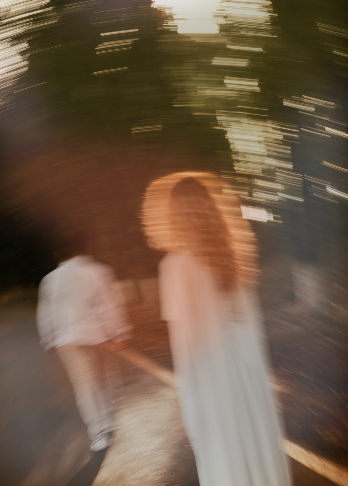 Blurred photo of a person with long red hair wearing a white dress, standing outdoors near a fence, at sunset or dusk.