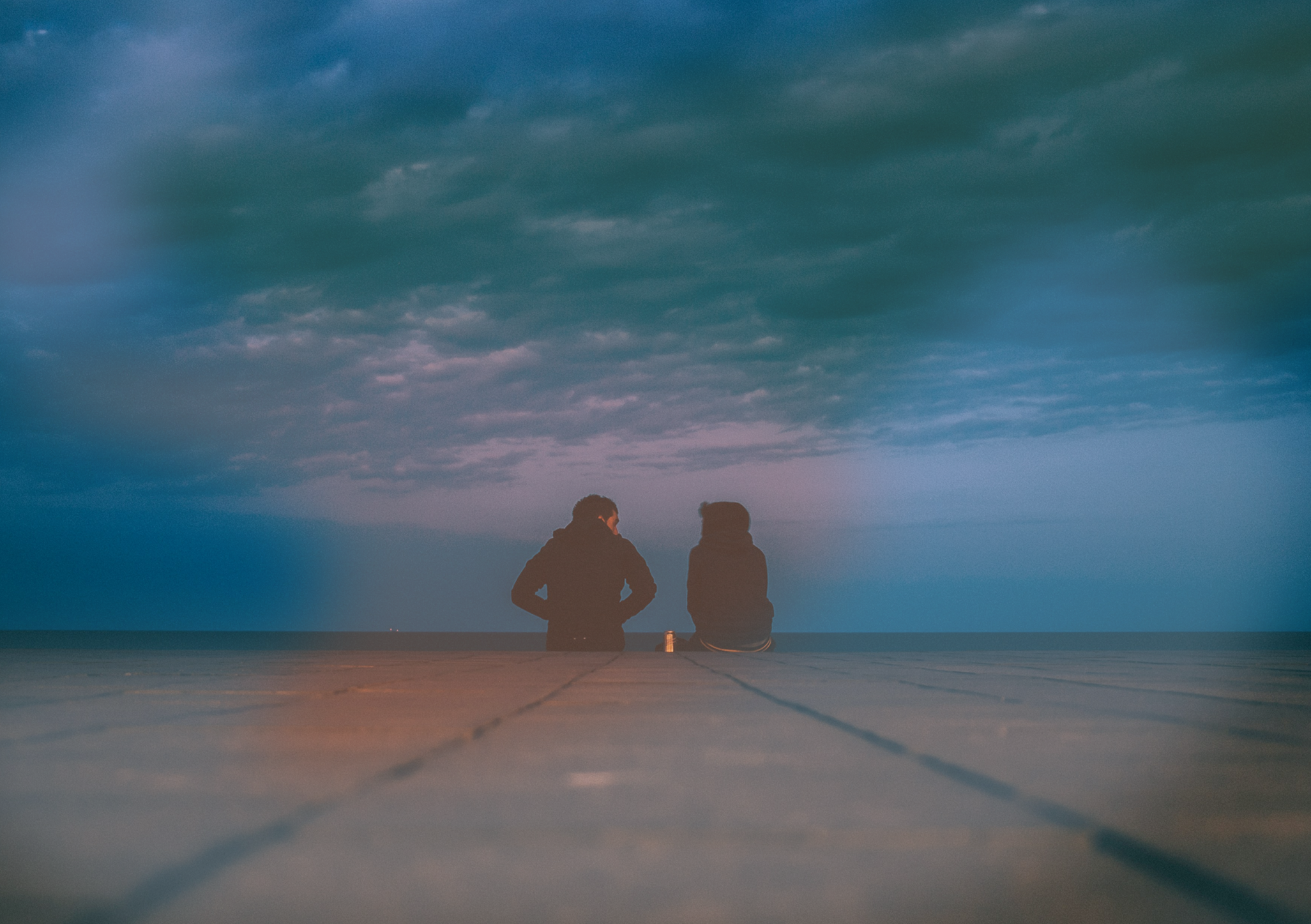 Two people sitting on a wooden dock at the beach during dusk, looking out over the ocean with a cloudy sky overhead.