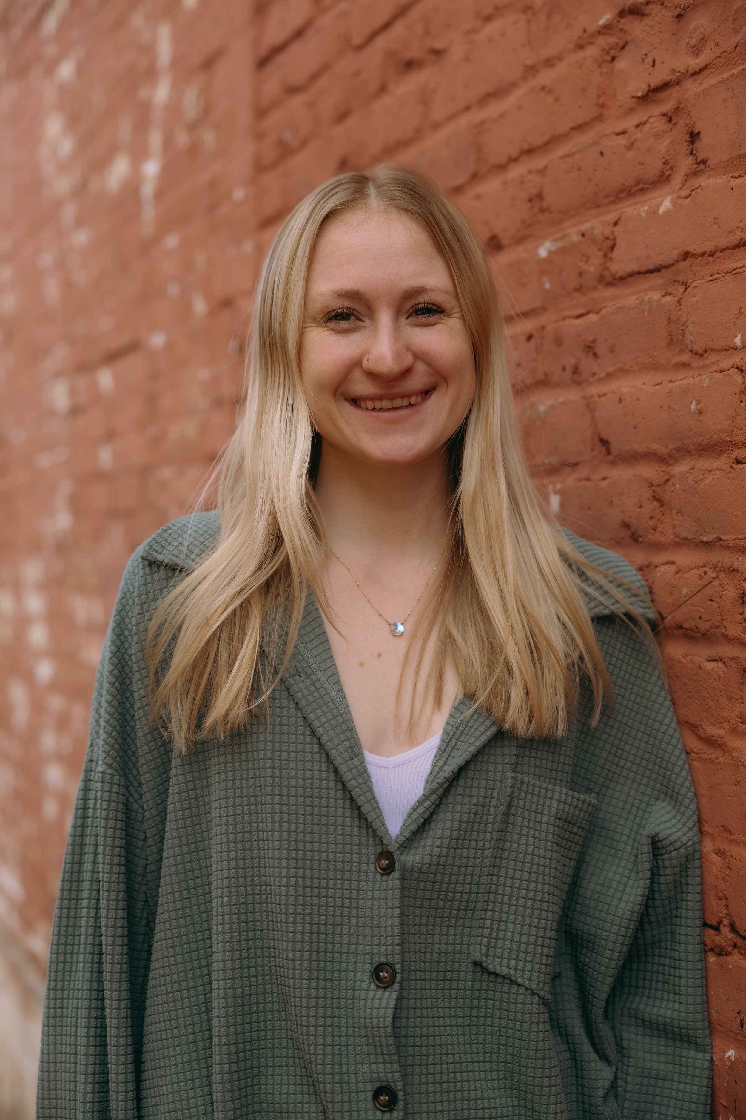 A young woman with blonde hair smiling, wearing a green checkered shirt over a white top, standing against a red brick wall.
