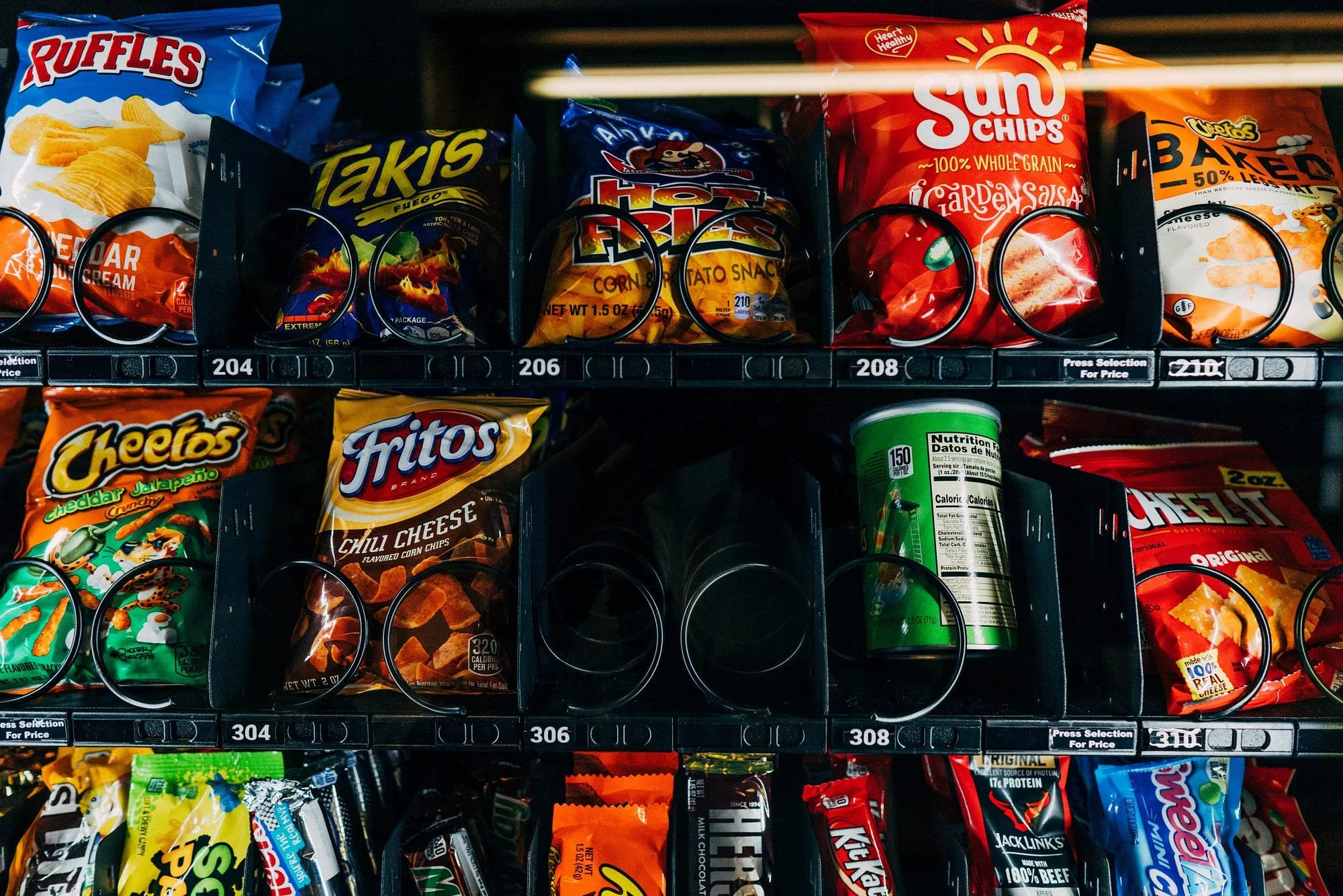 Vending machine snack aisle with various bags of chips and snacks, including Ruffles, Takis, Hot Fries, SunChips, Cheetos, Fritos, and other snack brands. Vending slots labeled with various prices.
