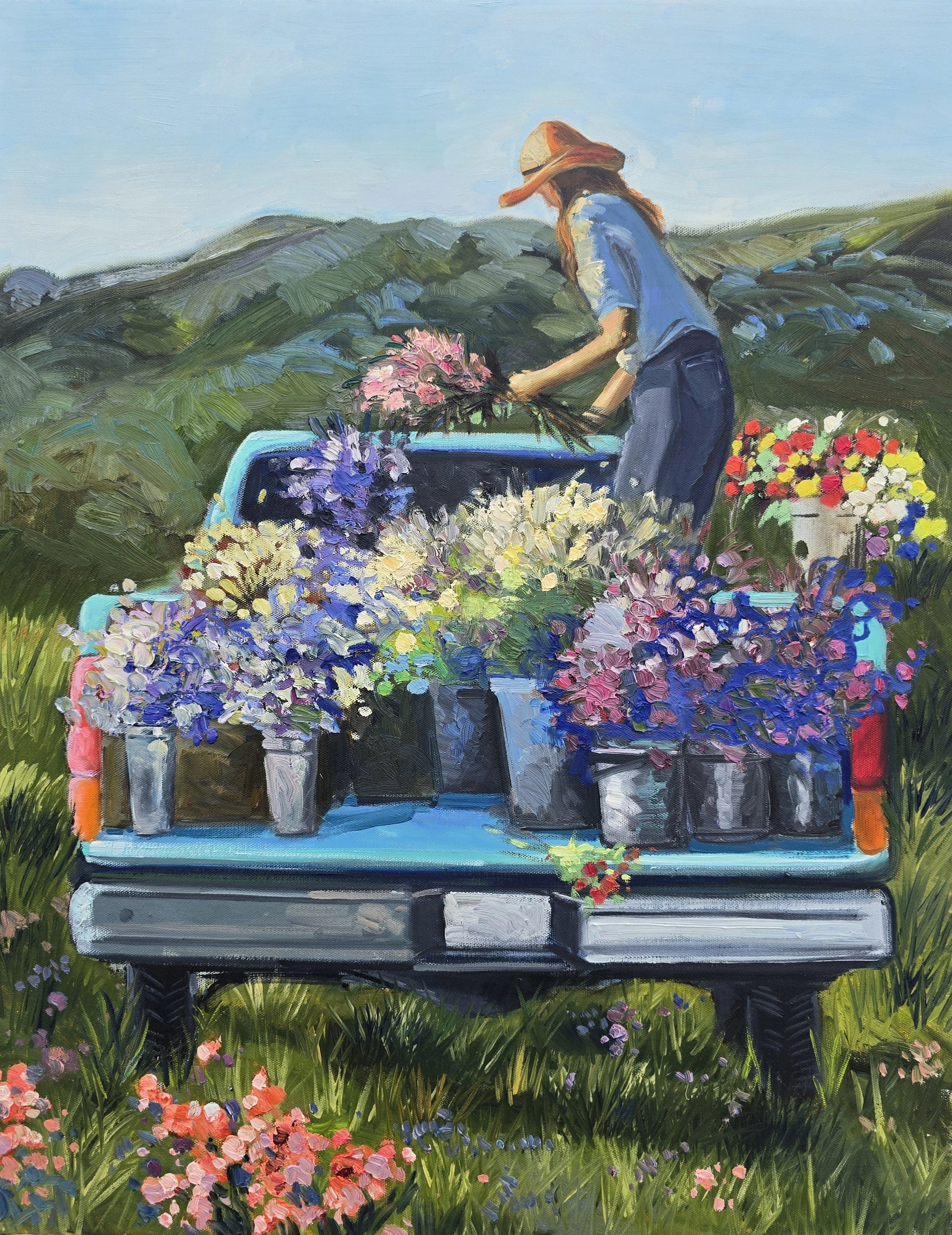 A woman in a straw hat and blue shirt arranging flowers in baskets on a table outdoors in a field of grass and wildflowers, with green hills and a blue sky in the background.