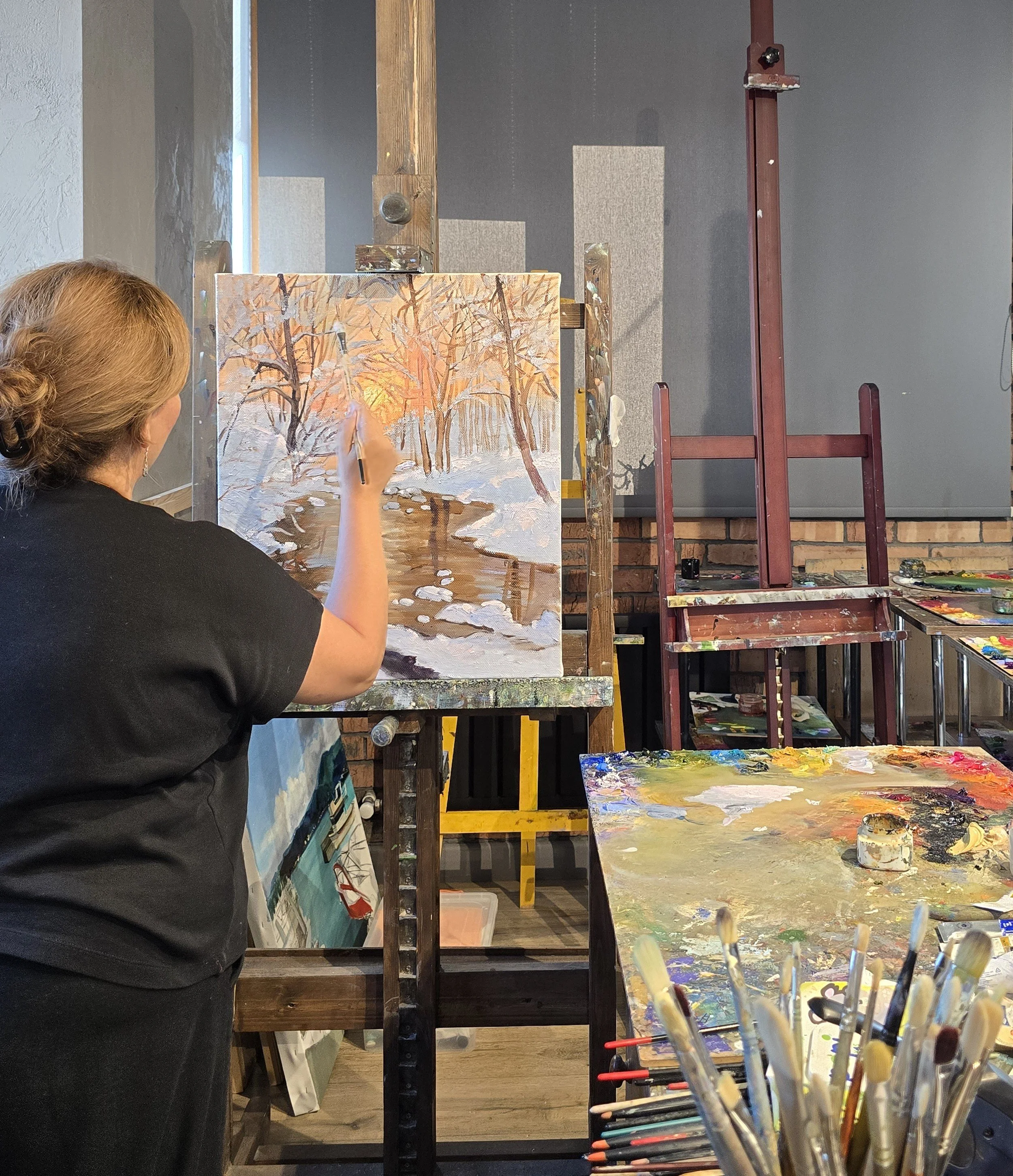 An artist painting a snow-covered landscape with a stream and trees with orange leaves on a canvas in an art studio.