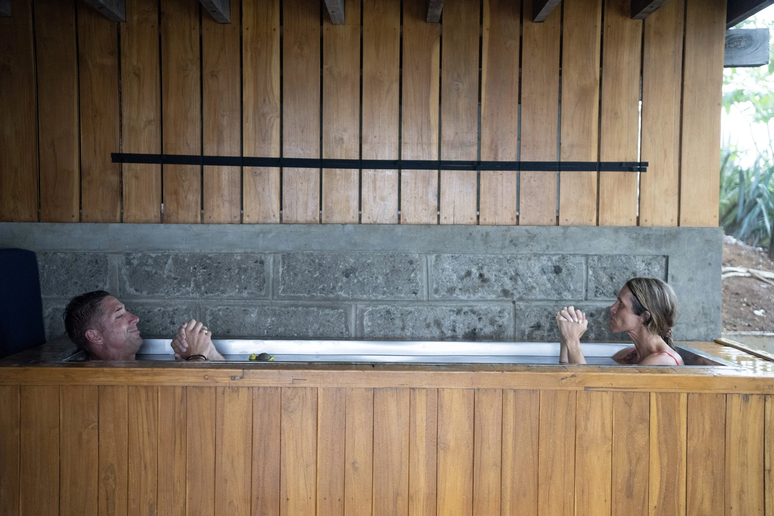 Two people, a man and a woman, are relaxing in an outdoor cold plunge with a wooden wall behind them. They are facing each other and engaging in meditation.