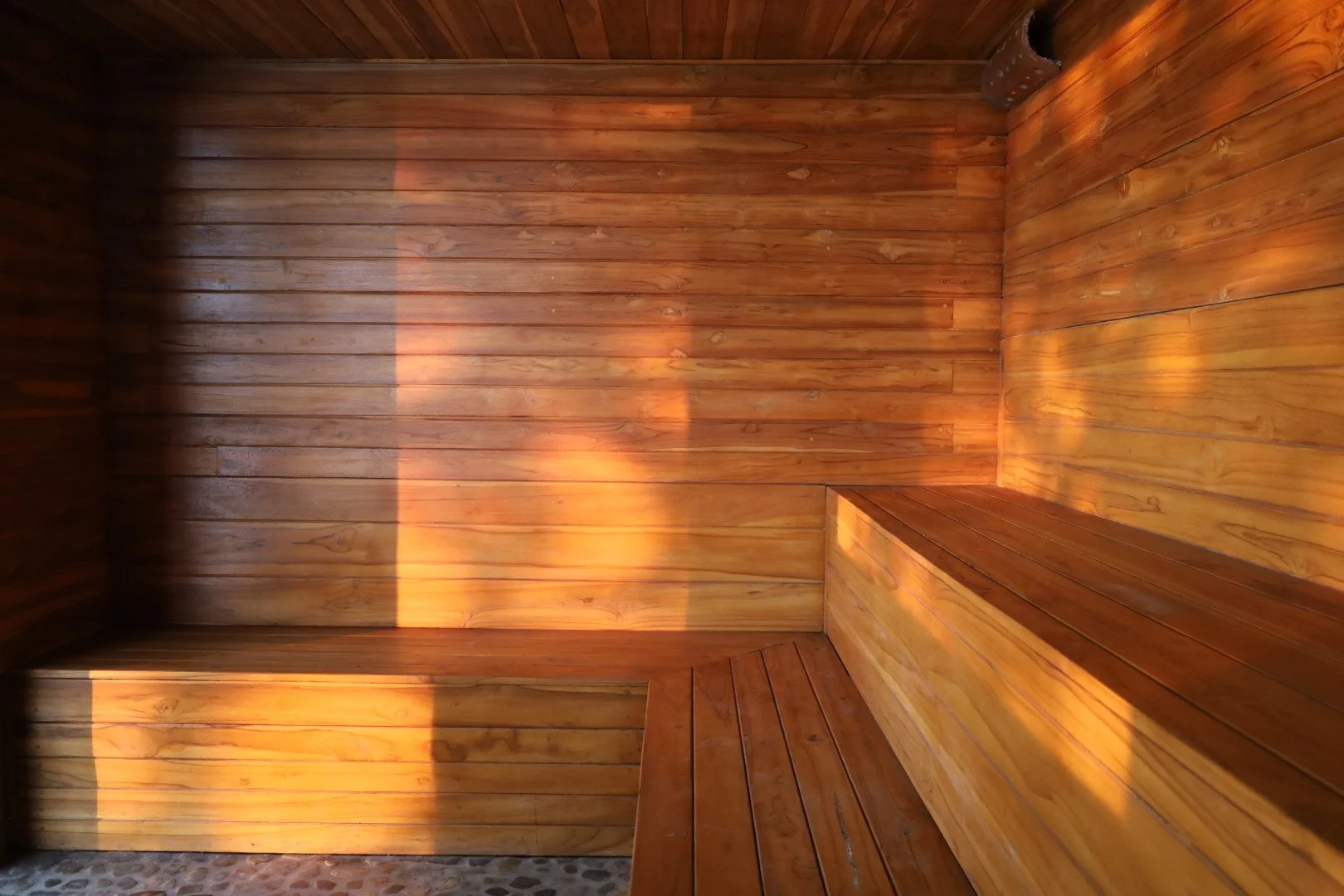 Interior of a wooden sauna with benches on the right side and sunlight casting shadow patterns on the wall.