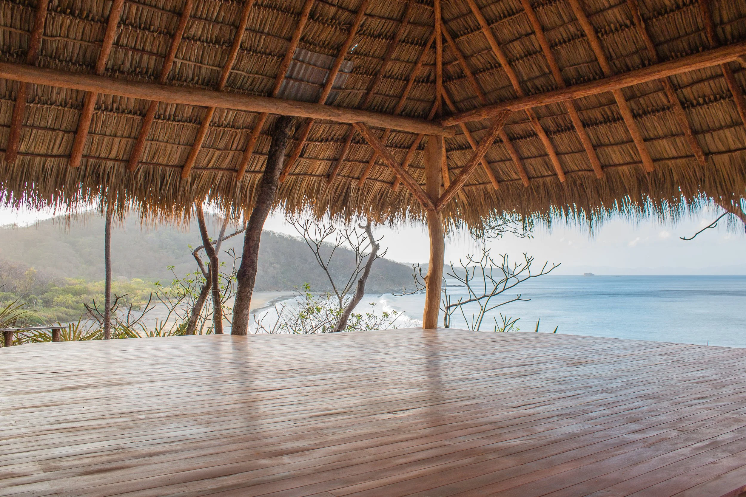 View from a yoga platform under a thatched roof hut overlooking a coastal landscape with trees, hills, and the ocean in the distance.