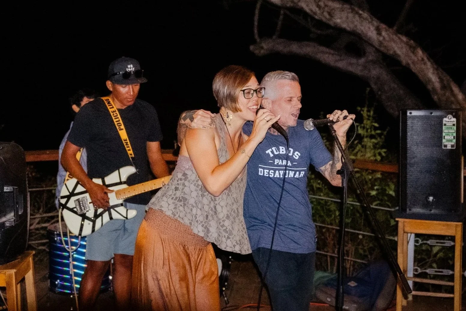 Two musicians singing into microphones while a guitarist plays in the background at an outdoor night event.