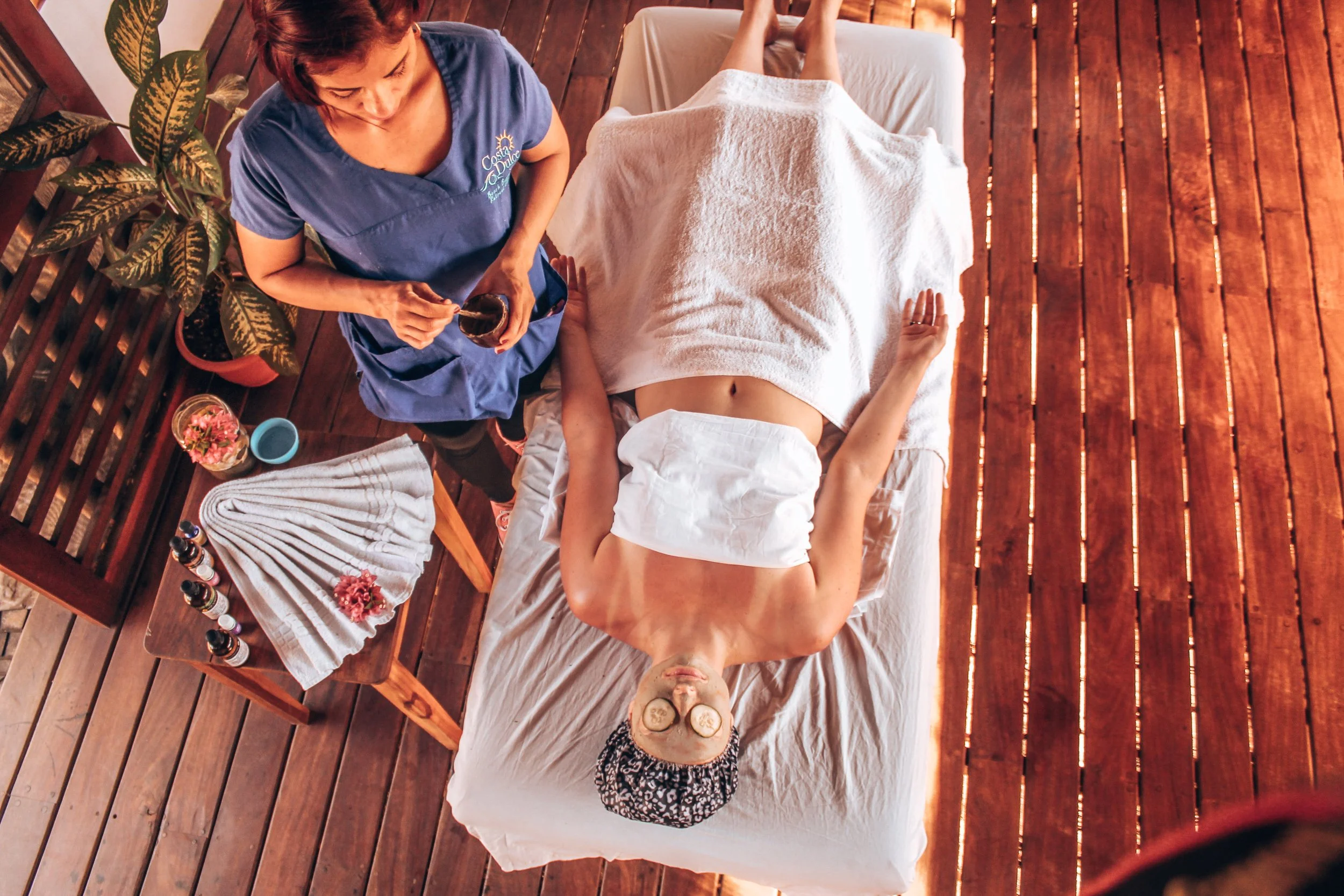 A woman receiving a facial massage on her back at a spa. The woman is lying on a massage table with a towel covering her lower body. A therapist wearing blue scrubs is standing next to her, holding a cup, possibly applying a treatment. The room has wooden flooring, and there is a small table with skincare products, a towel, and flowers nearby.