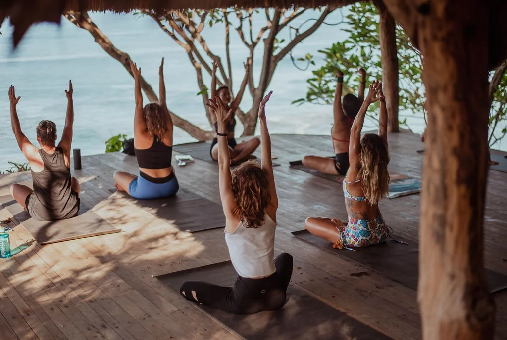 Six women practicing yoga outdoors on a wooden deck near water, sitting cross-legged with arms raised in a yoga pose, under a thatched roof with trees and water in the background.
