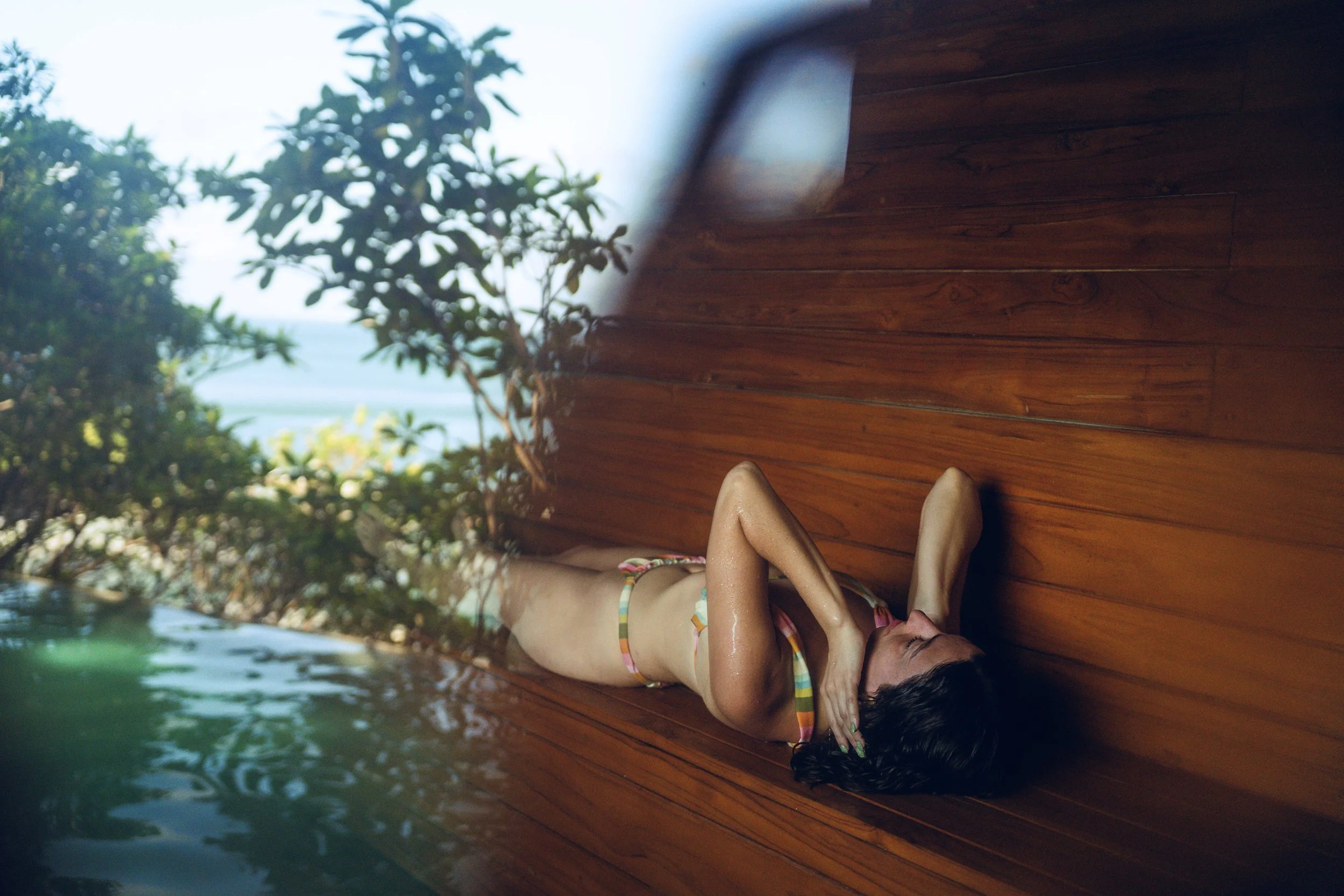 A woman in a bikini lying in a sauna, poolside deck with trees and water is reflected in the sauna window.