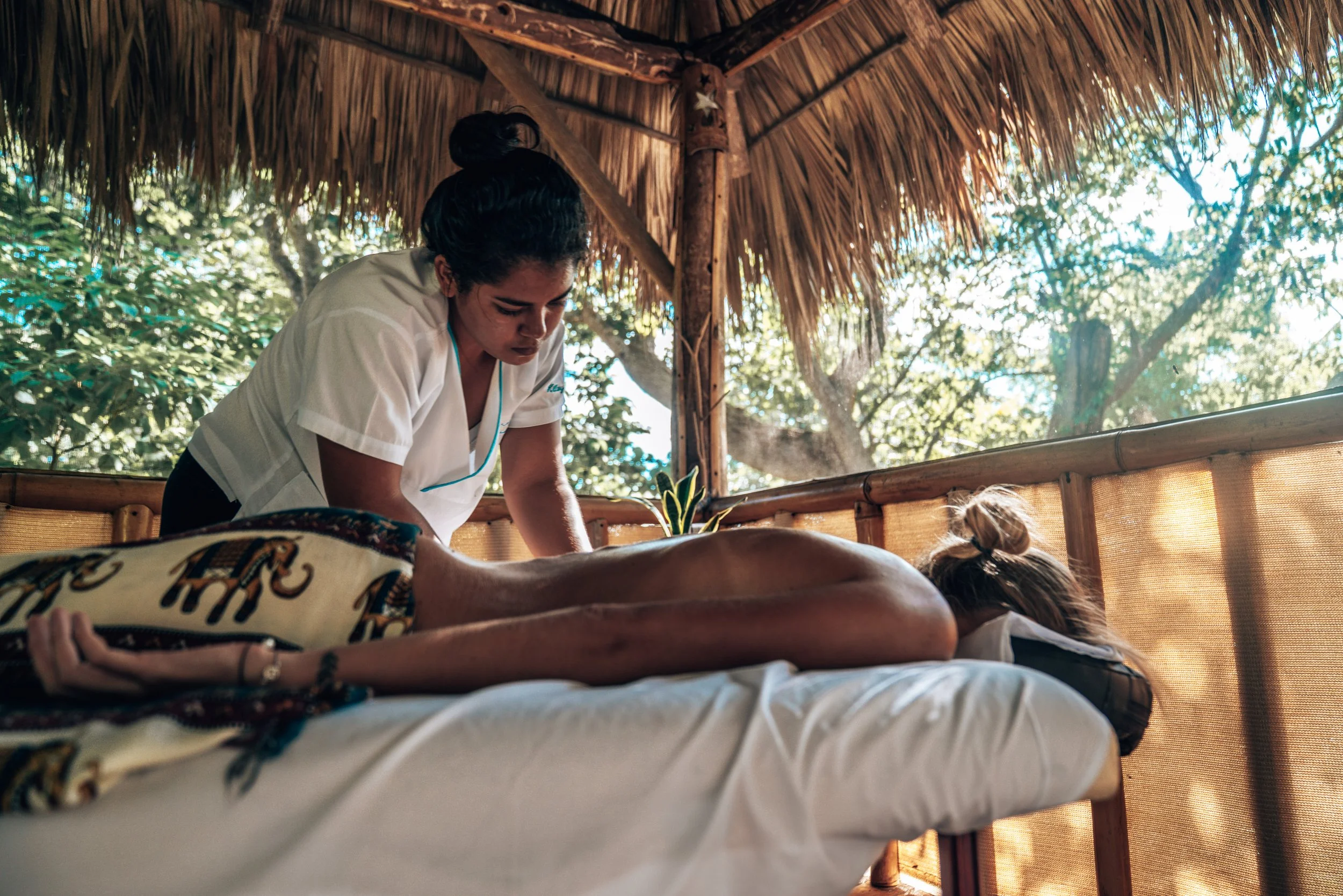 A woman receiving a massage outdoors under a thatched roof surrounded by trees.