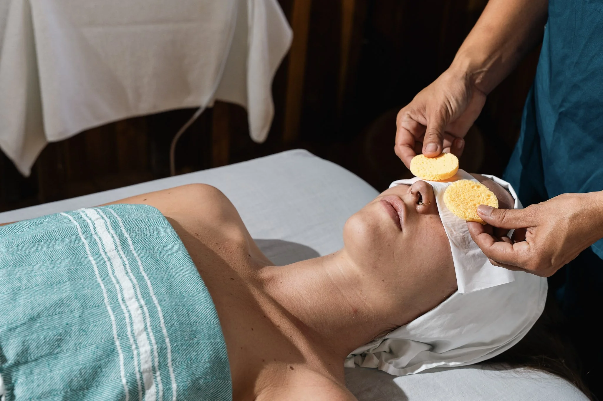 Person lying on a massage table, receiving a facial with a sponge, while another person applies a facial treatment to their face with sponge.
