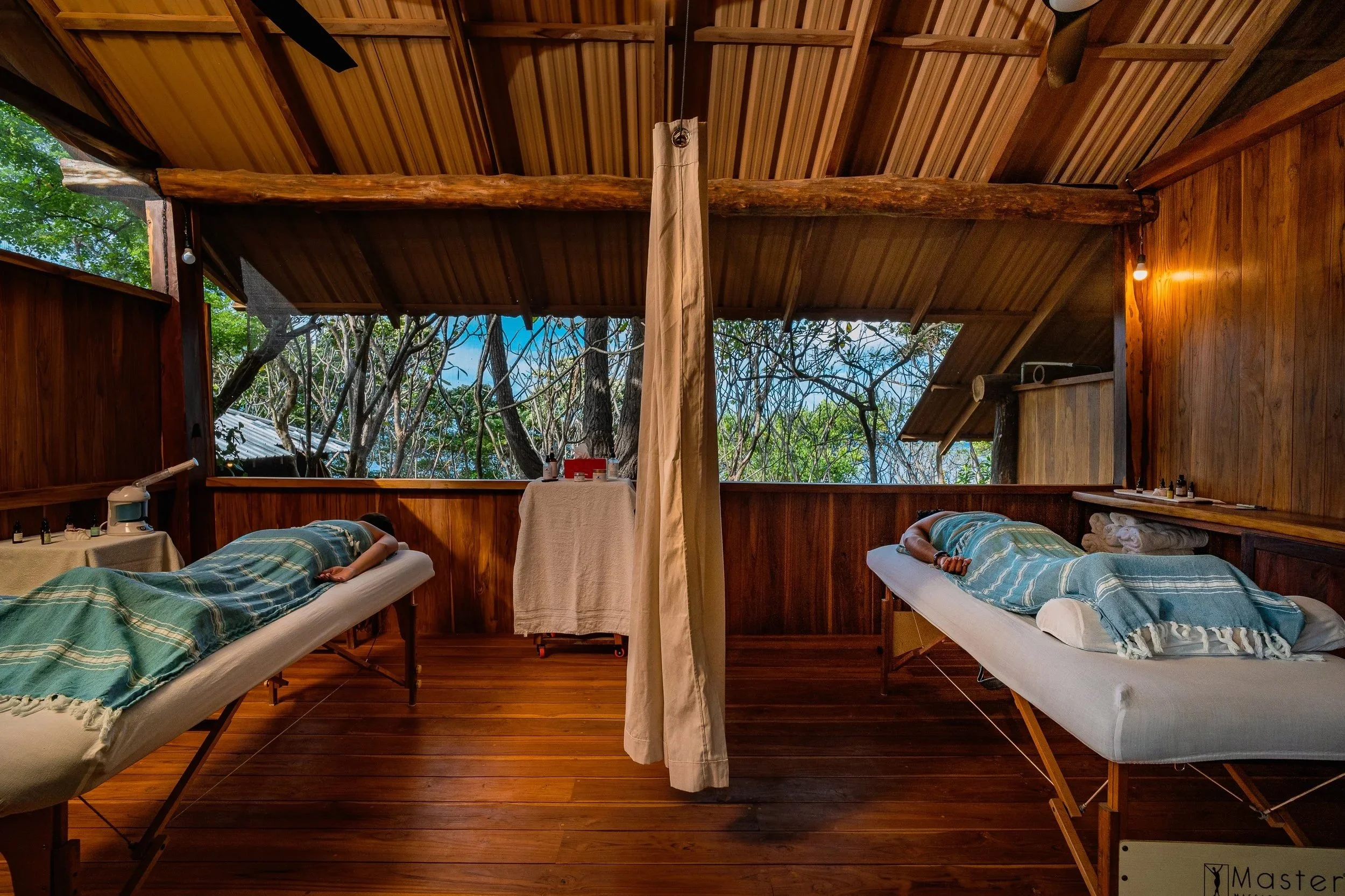 Two massage tables with person lying face down, in a wooden room with open window showing trees outside, and soft lighting.