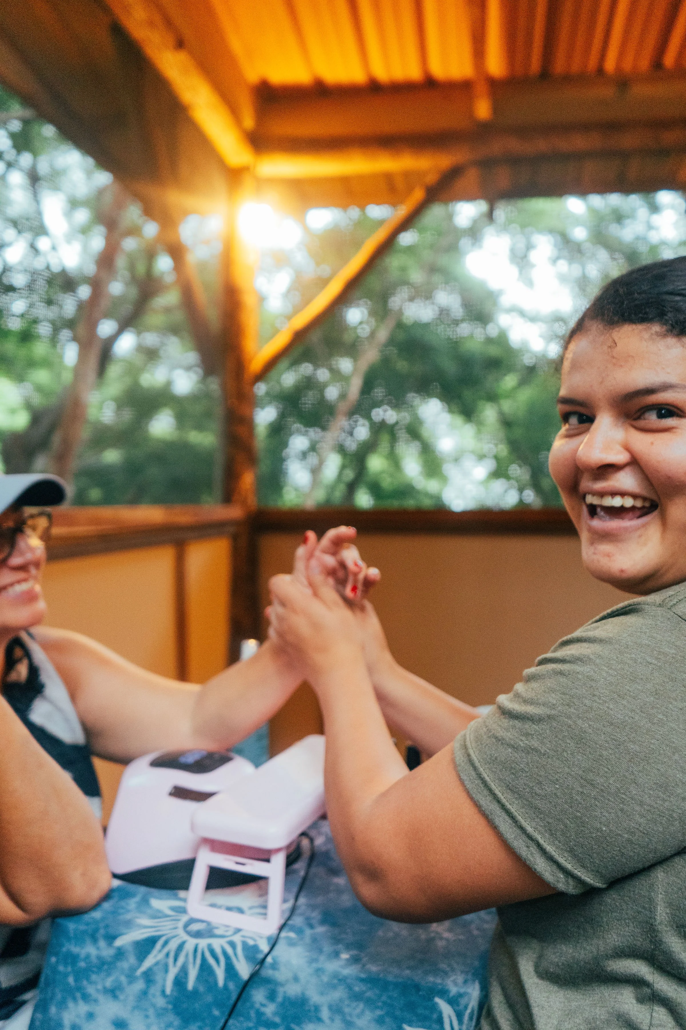 A woman gives a manicure in a massage cabin with trees in the background, sunlight shining through.
