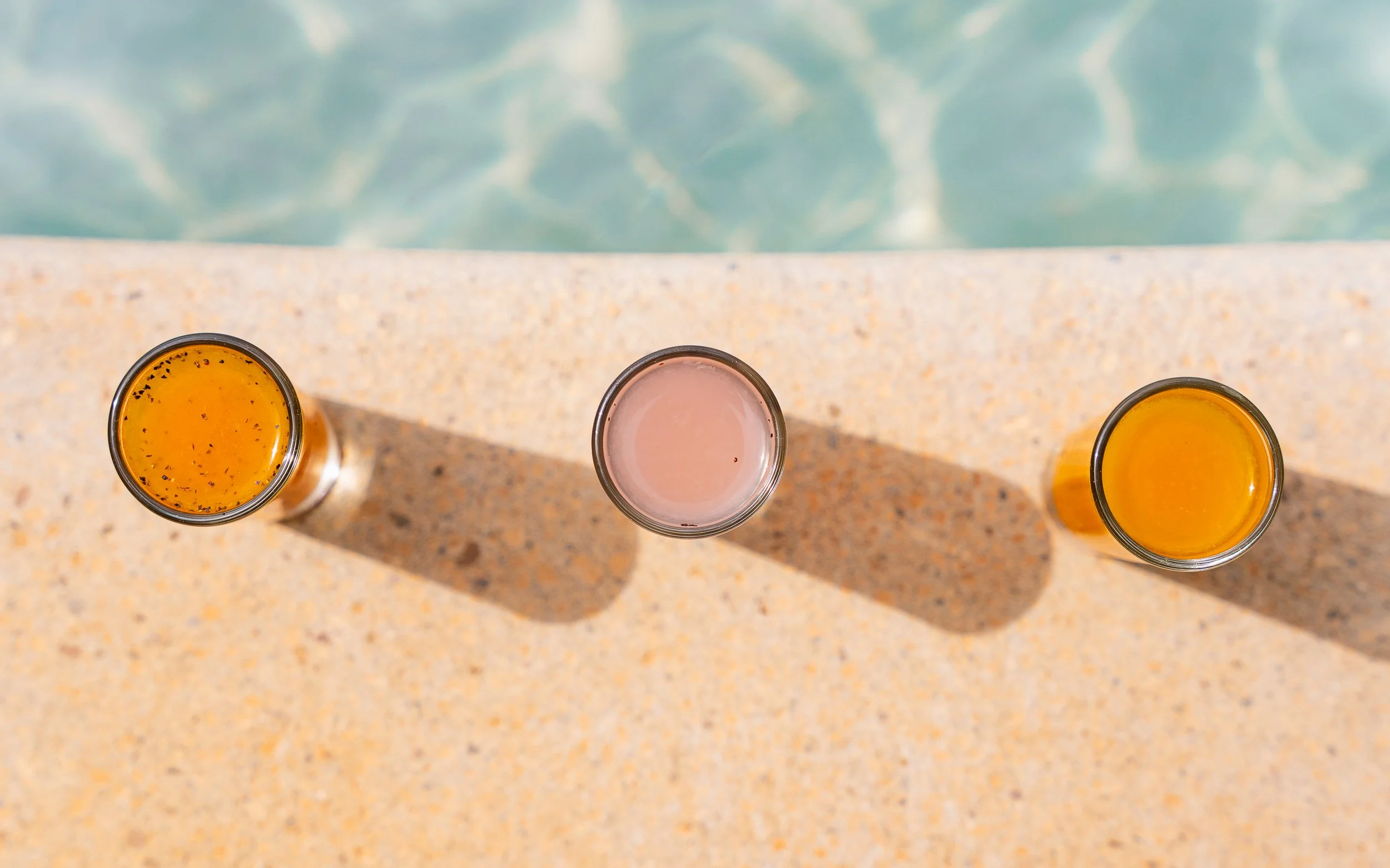 Three glasses filled with orange, pink, and orange beverages are placed on a speckled outdoor table near a swimming pool. The top-down view captures the glasses and their shadows.