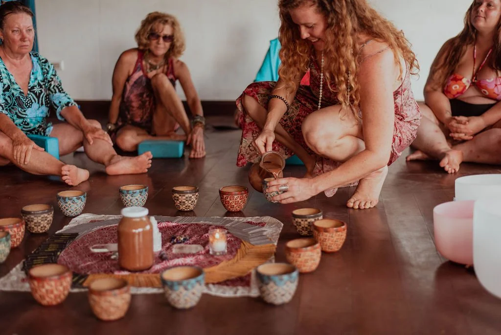 A group of women sitting on the floor in a circle, with a woman in the center pouring a cup during a meditation or spiritual gathering with candles and decorated cups on the floor.