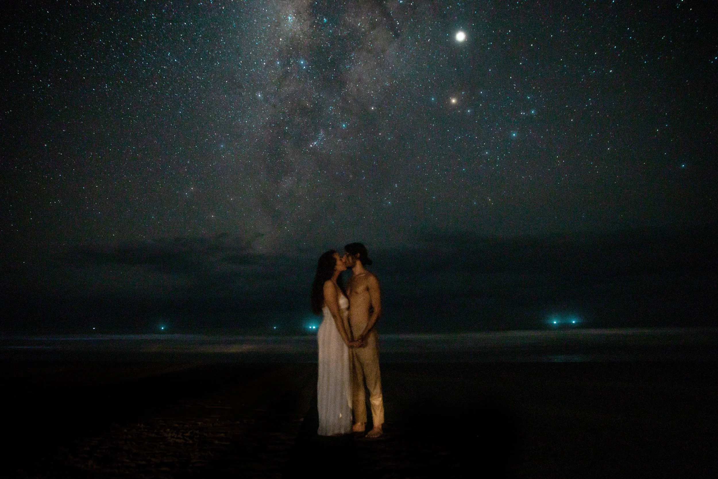 A couple stands on a beach at night, kissing under a starry sky with the Milky Way visible.