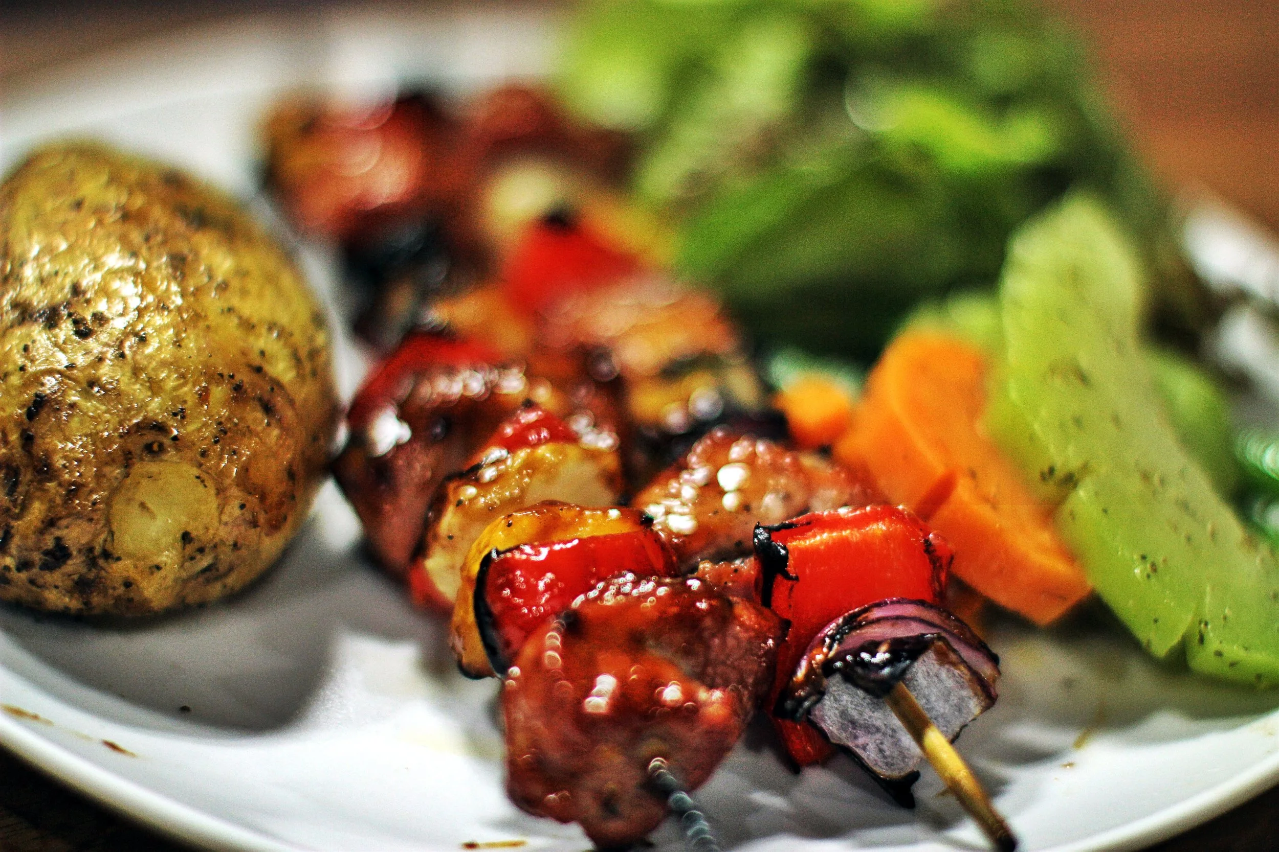 Close-up of grilled vegetable skewers on a white plate, including red bell peppers, purple onions, and yellow squash. In the background, there's a baked potato and some green salad with orange carrots.