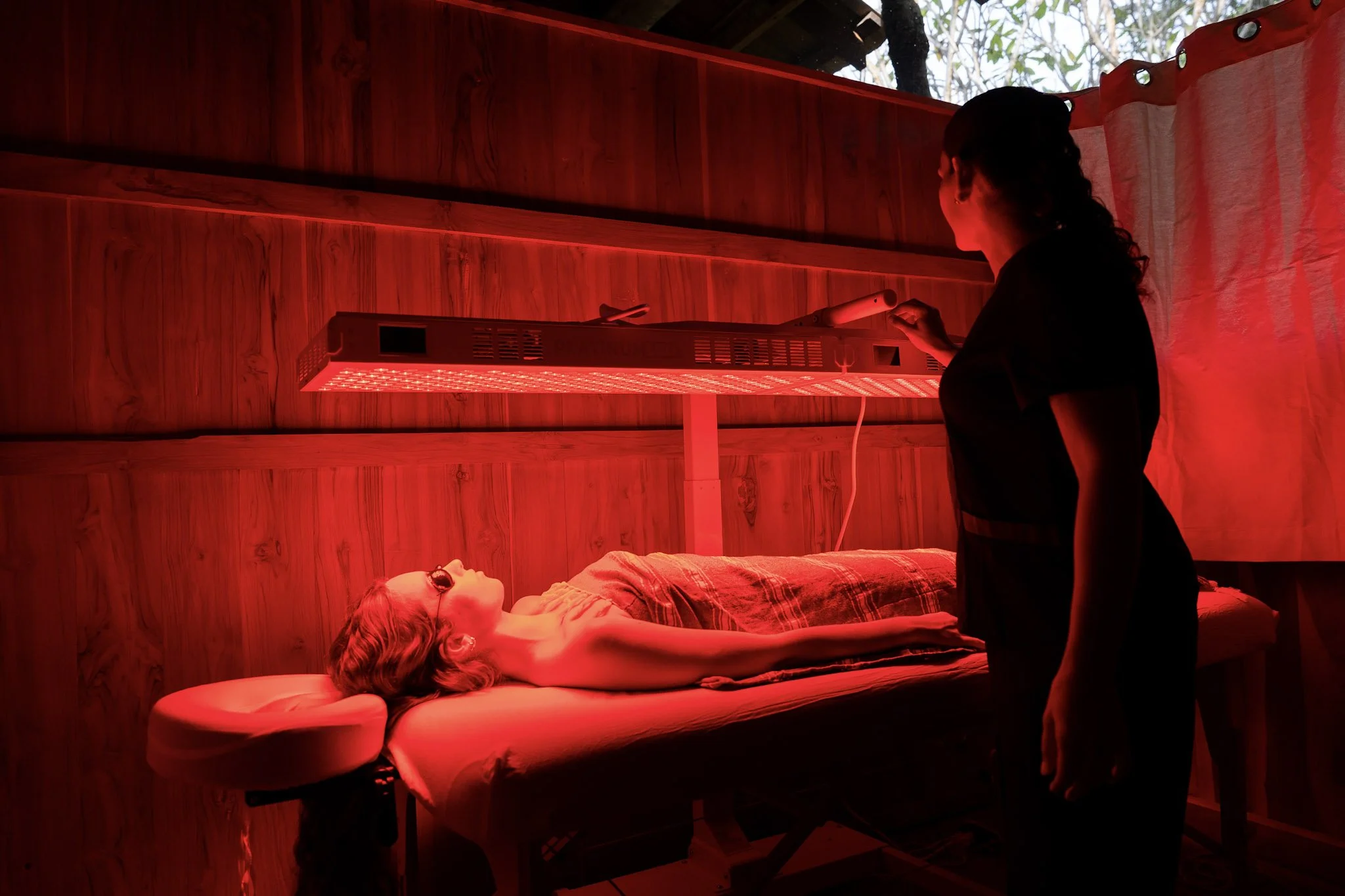 A woman lies on a massage table with headphones on in a room illuminated by red light. A therapist stands nearby, holding a device, in front of a wooden wall with a window showing trees outside.