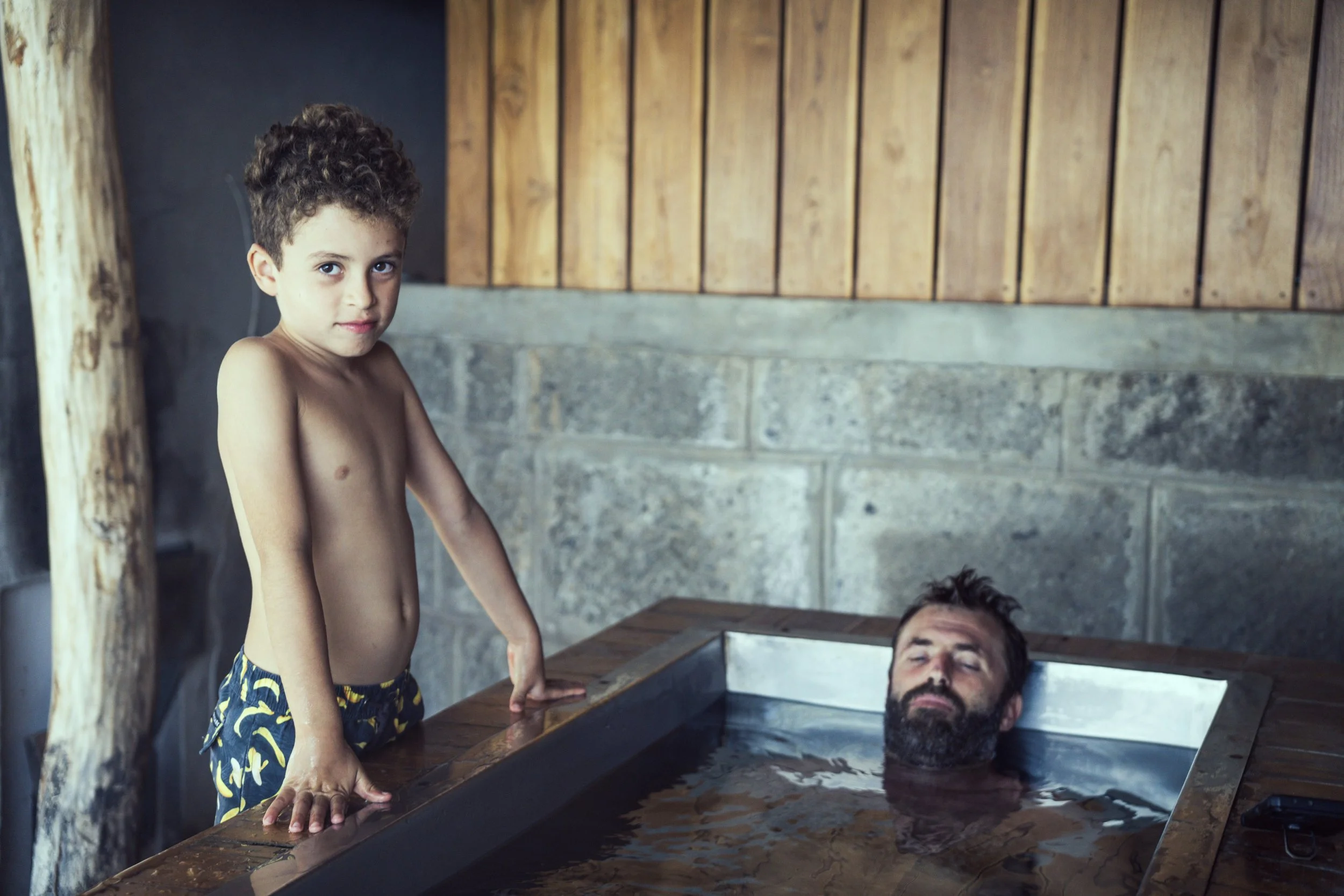 A young boy with curly hair standing next to a cold plunge, while an adult man with a beard relaxes in the cold plunge while meditating.