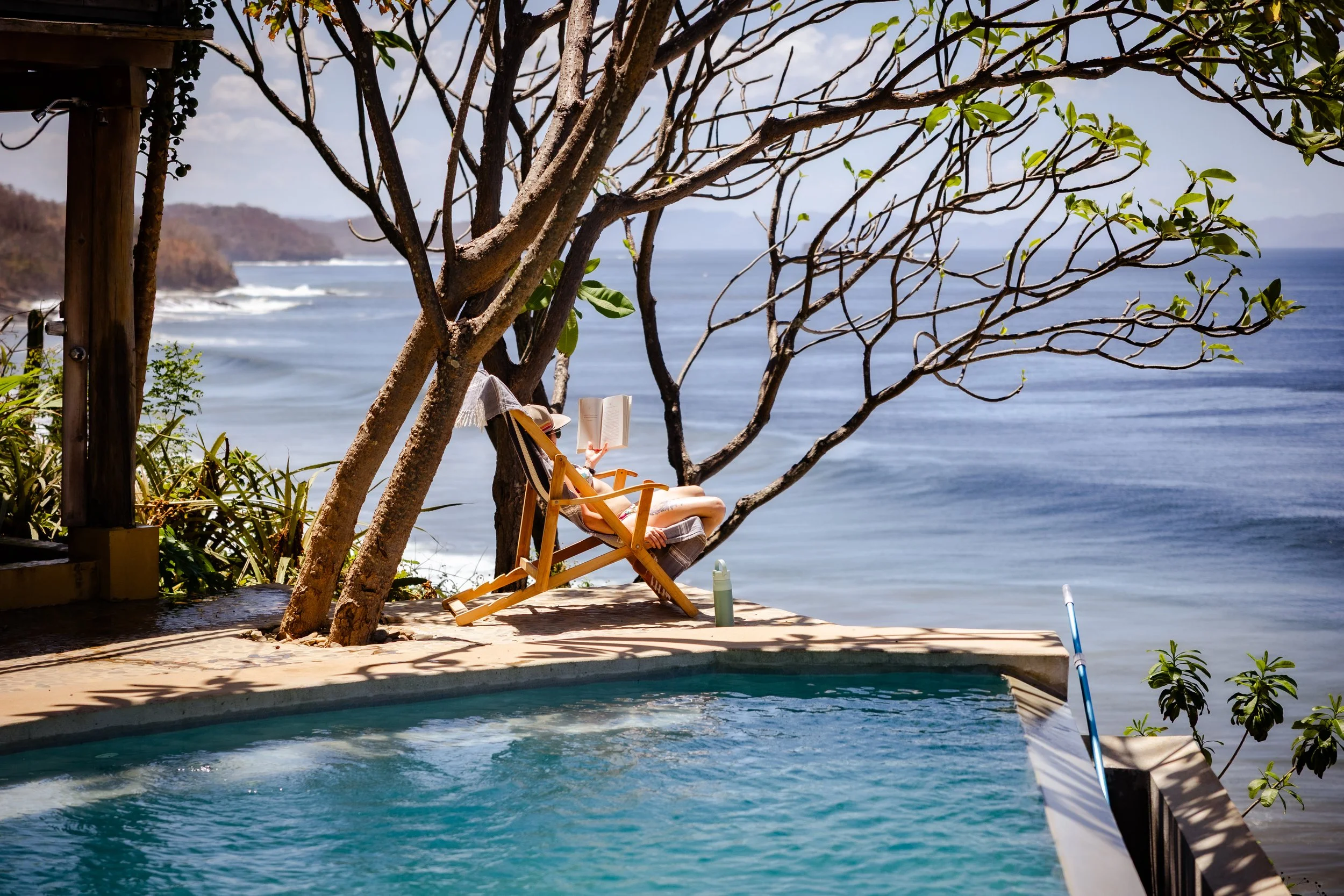 A person relaxing on a wooden beach chair reading a book under a tree near the ocean, with a view of cliffs in the distance, on a sunny day.