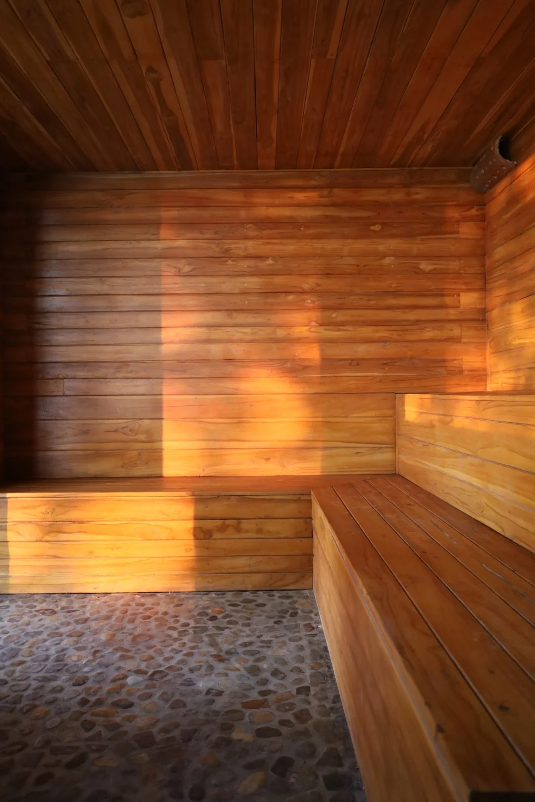 Wood-paneled sauna with pebble floor and sunlight streaming through a window.