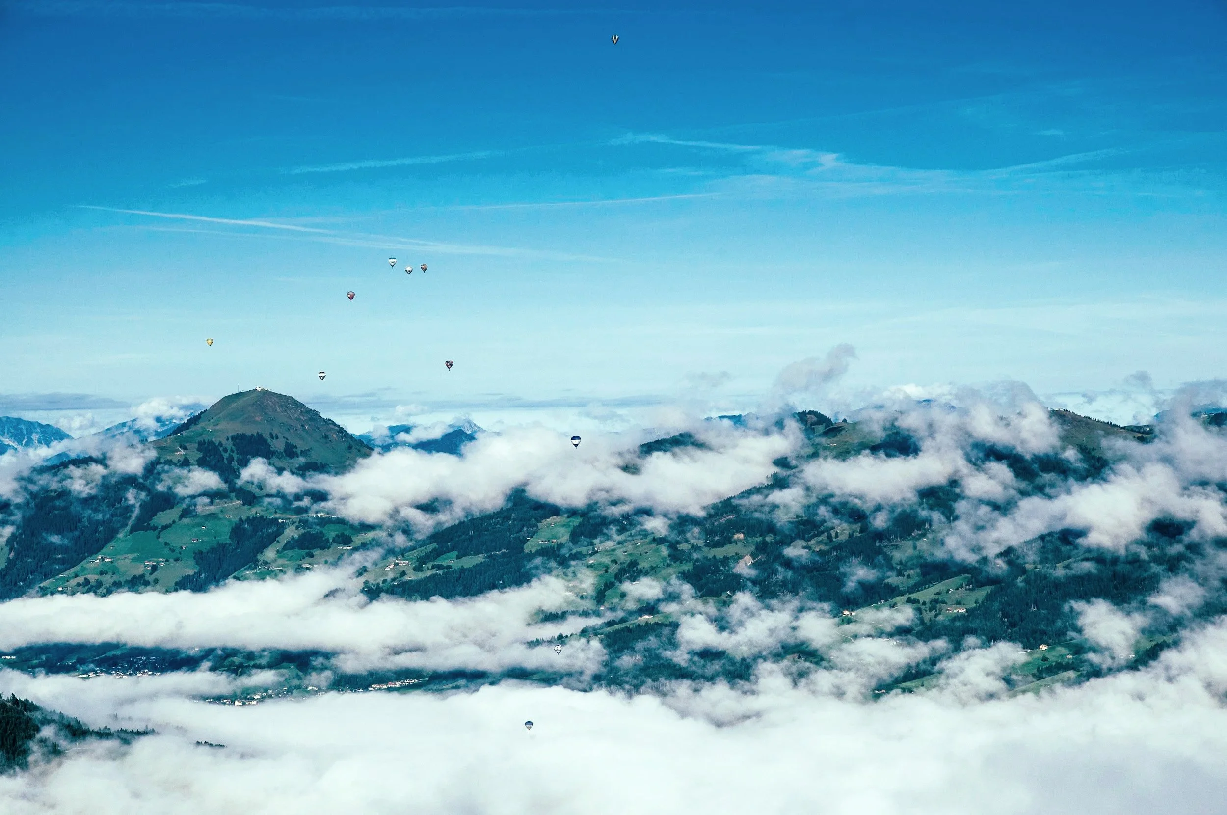 Luftaufnahme mit mehreren Heißluftballons über grün bewaldeten Hügeln, Wolken im Himmel.